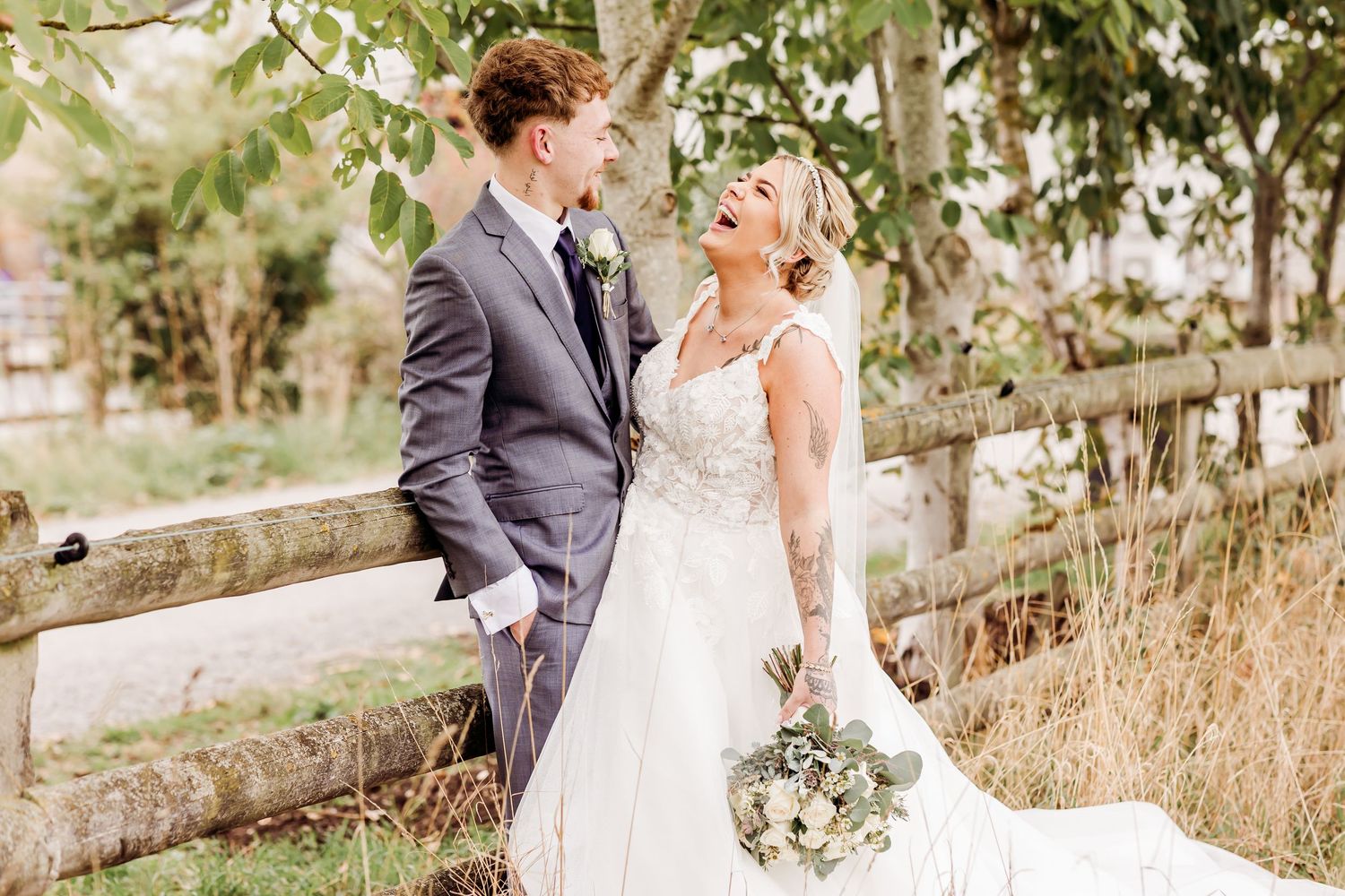 Bride and groom smiling at each other by rustic wooden fence, bride holding white bouquet in elegant lace wedding dress.