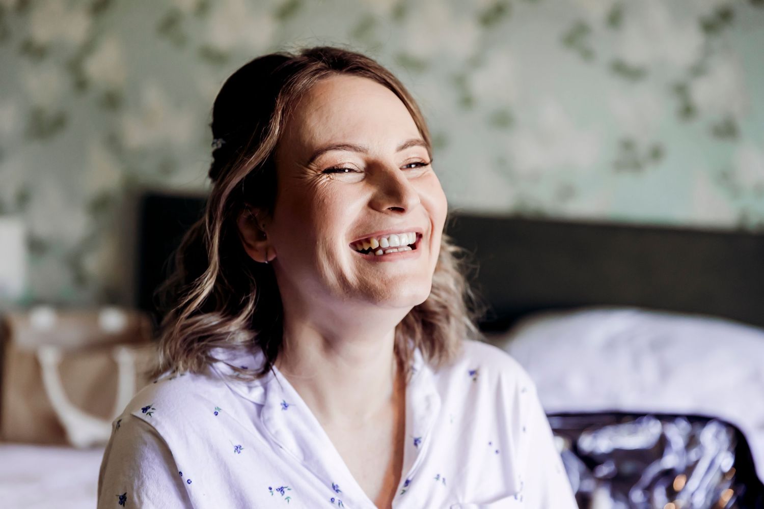 Smiling bride in floral pyjamas getting ready in a room with vintage wallpaper.