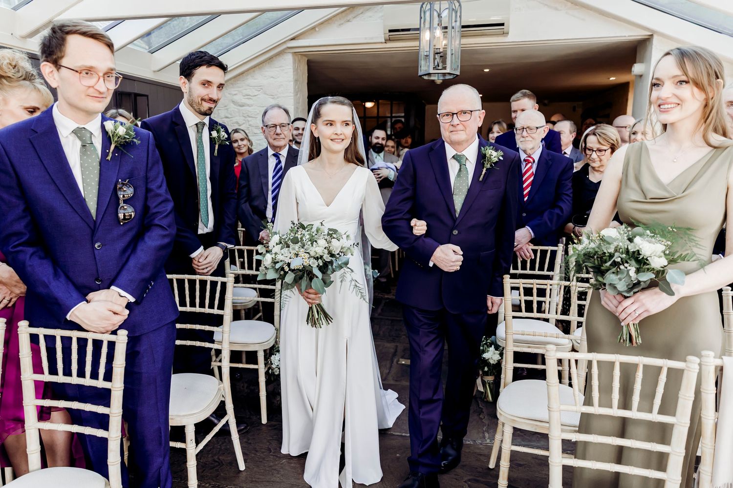Bride in a white jumpsuit walking down the aisle with her father holding a greenery bouquet.