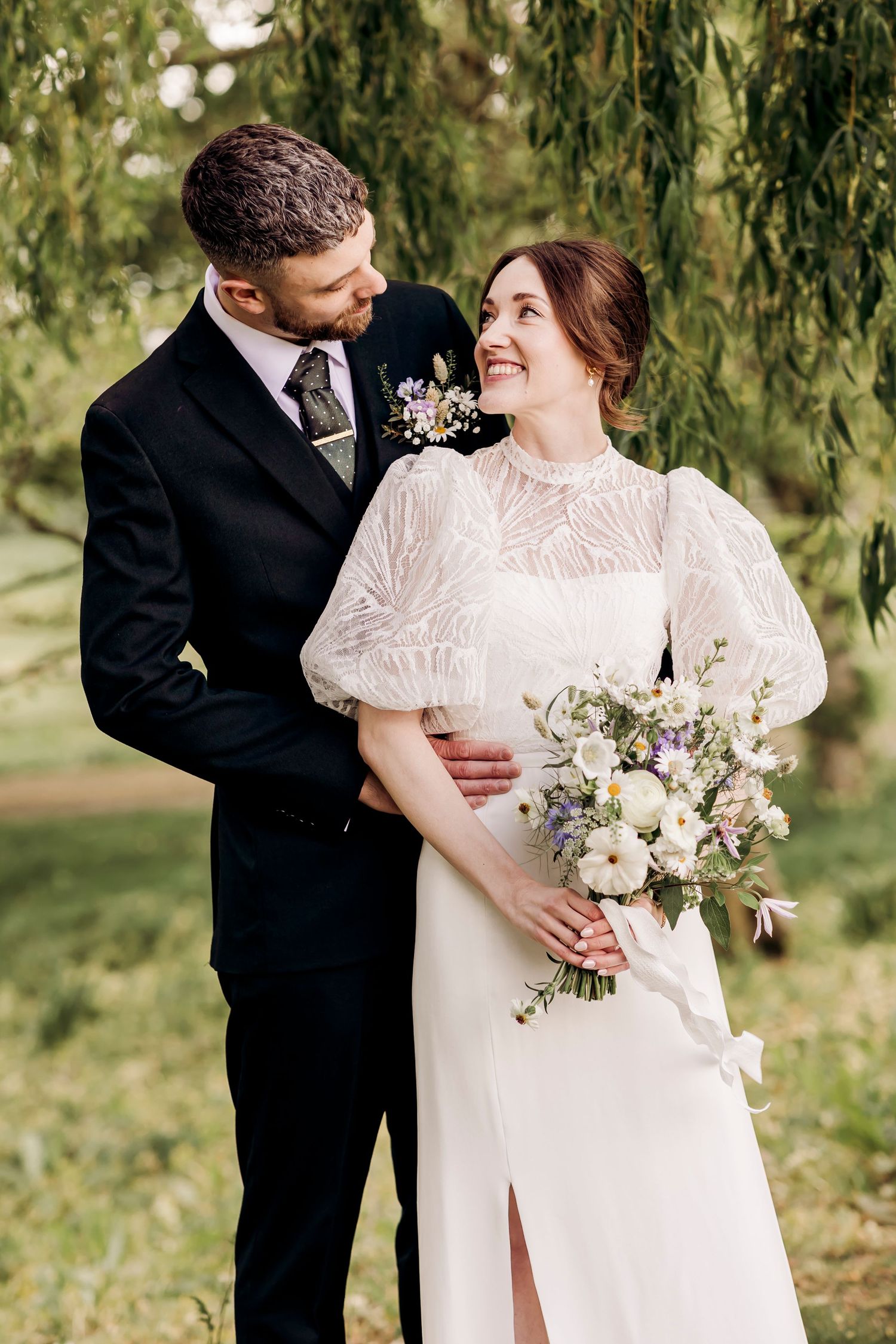 Bride and groom embrace outdoors under weeping willow tree, bride holding white and purple bouquet in elegant lace wedding dress.