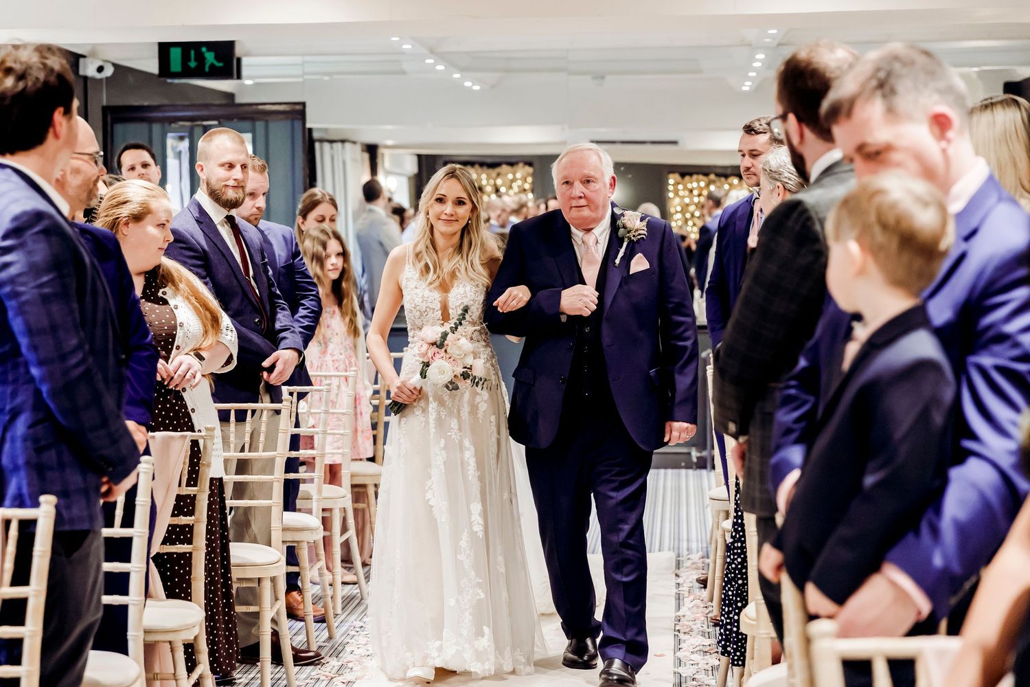 Bride in lace gown walking down aisle escorted by father at elegant wedding ceremony.