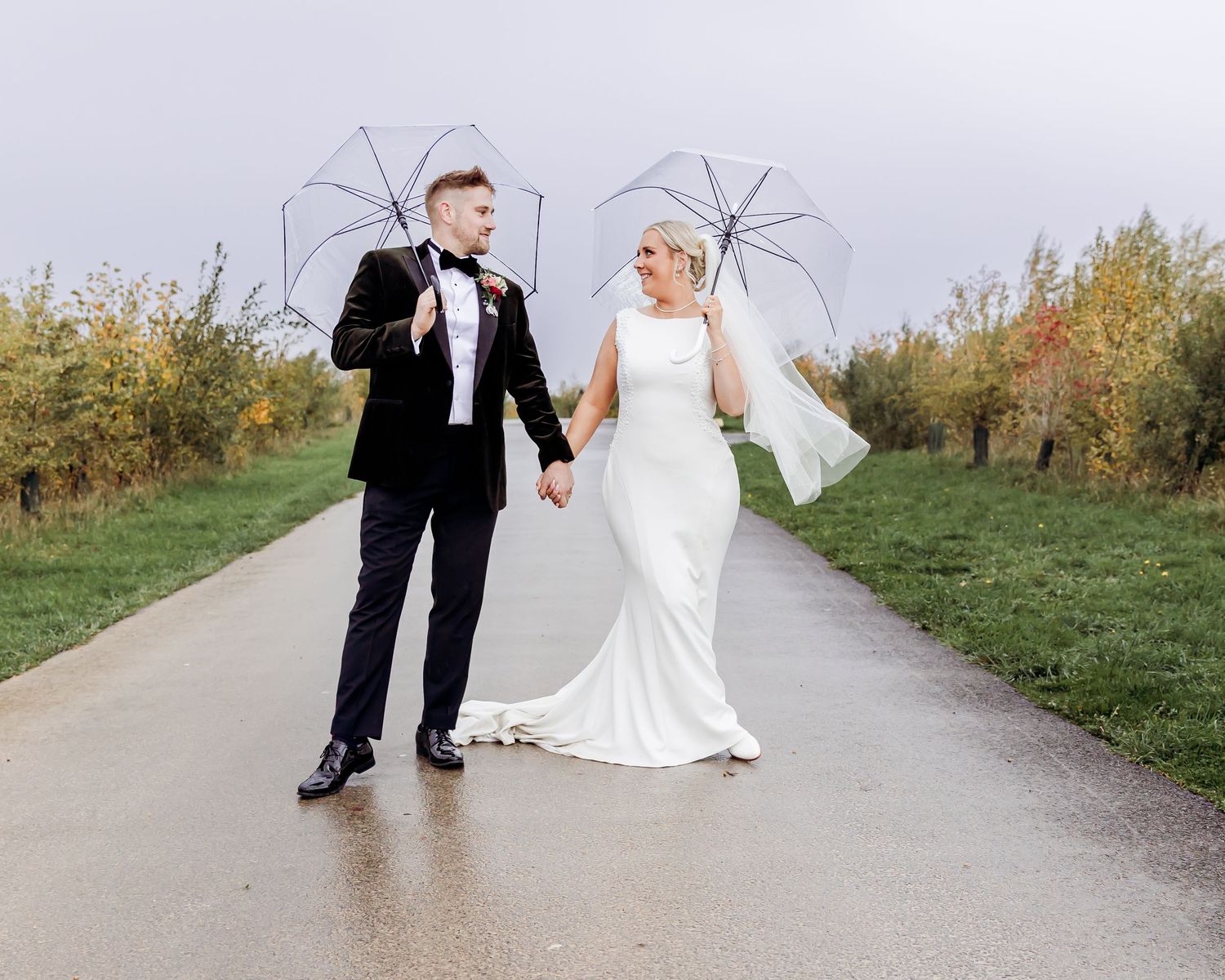 Bride and groom walk hand in hand under clear umbrellas on a rainy wedding day.