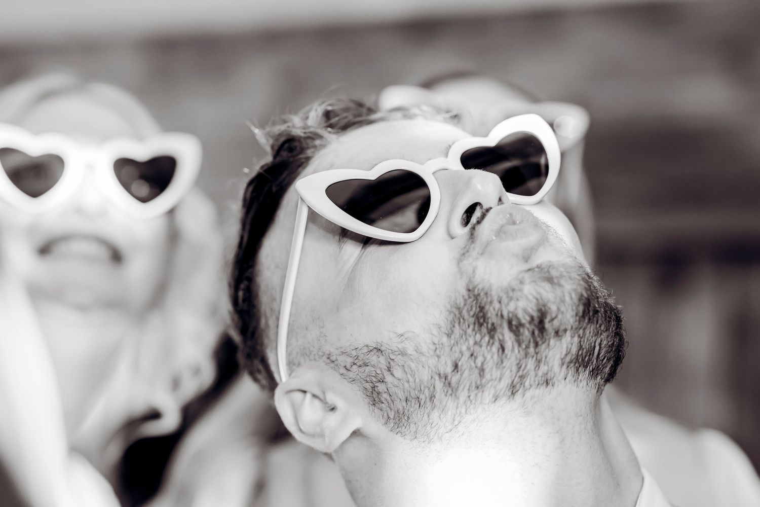 Black and white photo of bearded man wearing heart-shaped sunglasses at a fun wedding celebration.