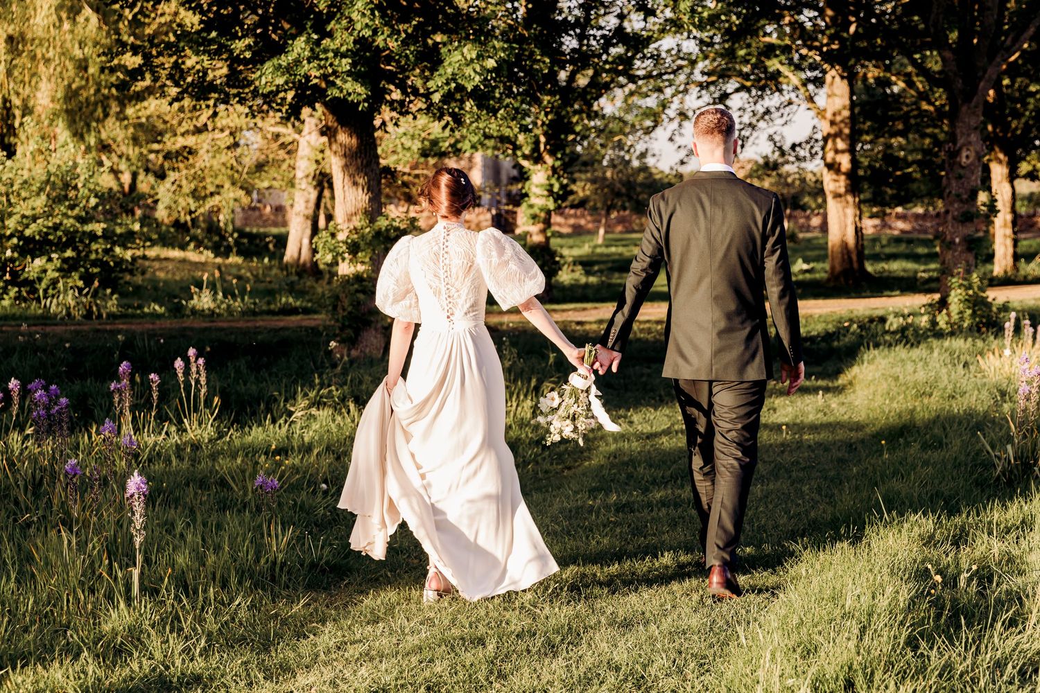 Bride and groom walking hand in hand through a lush green garden, with the bride holding a bouquet.