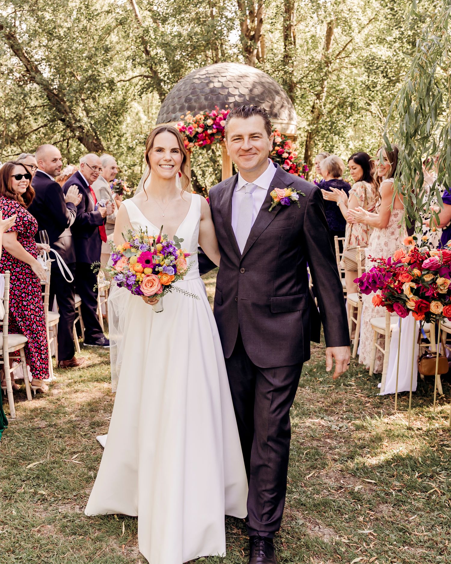Happy newlyweds walking down outdoor wedding aisle surrounded by colorful floral arrangements and guests.