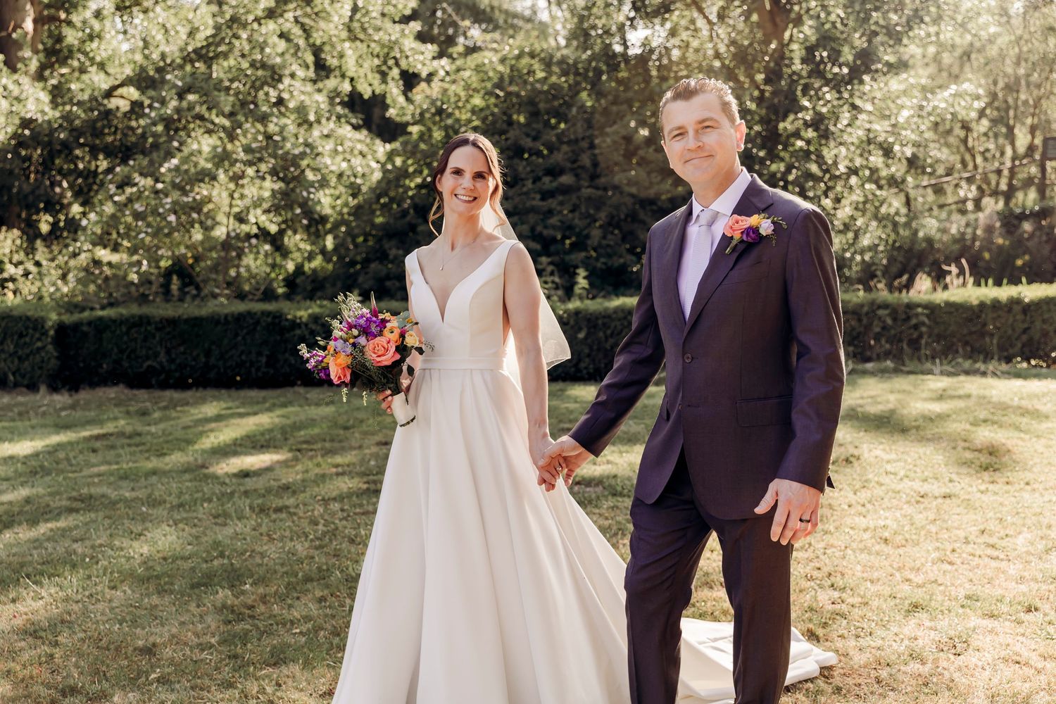 Bride in white gown holding colorful bouquet stands with groom in purple suit on sunny garden lawn.
