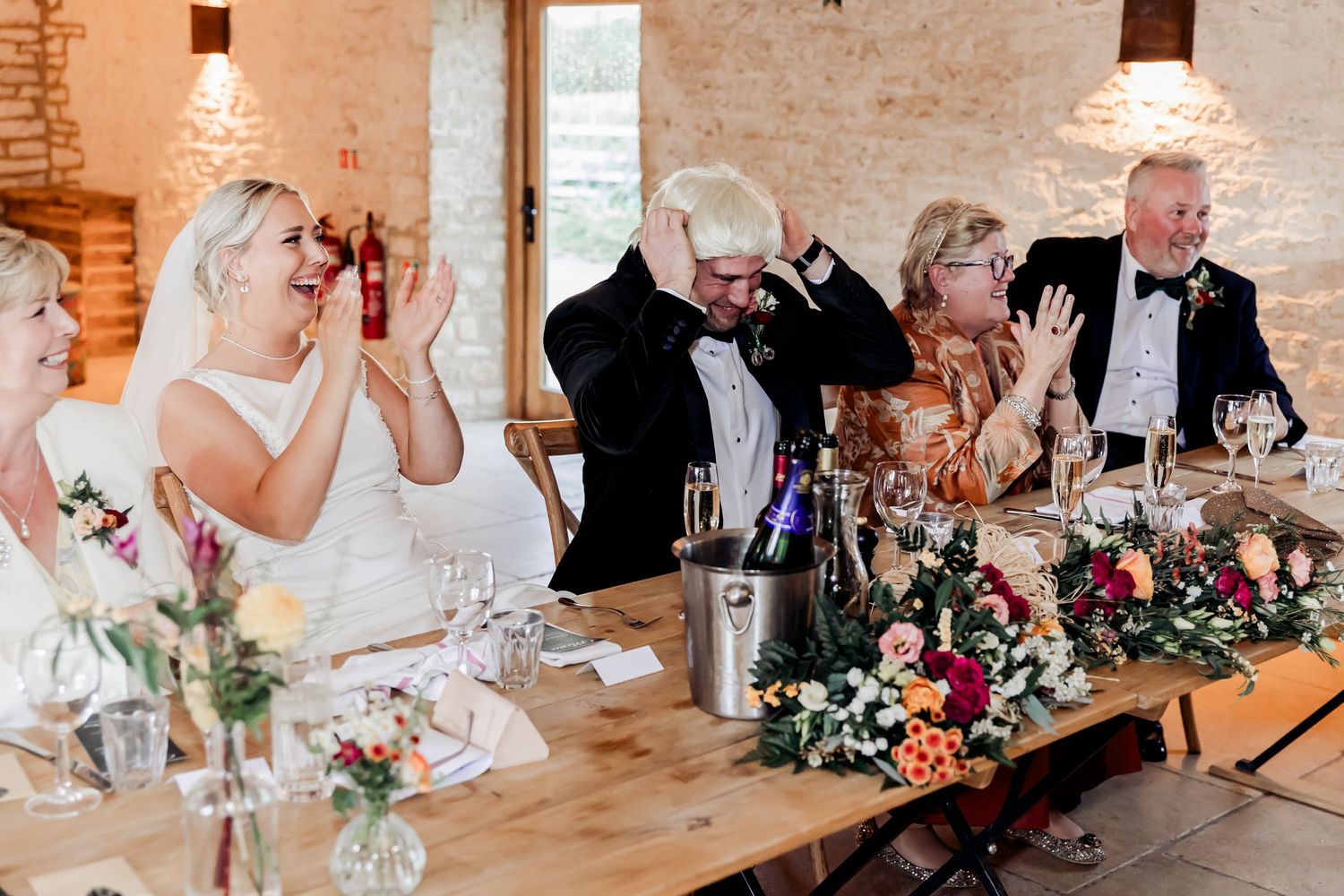 Bride and groom laugh with family at a floral-decorated wedding reception head table.
