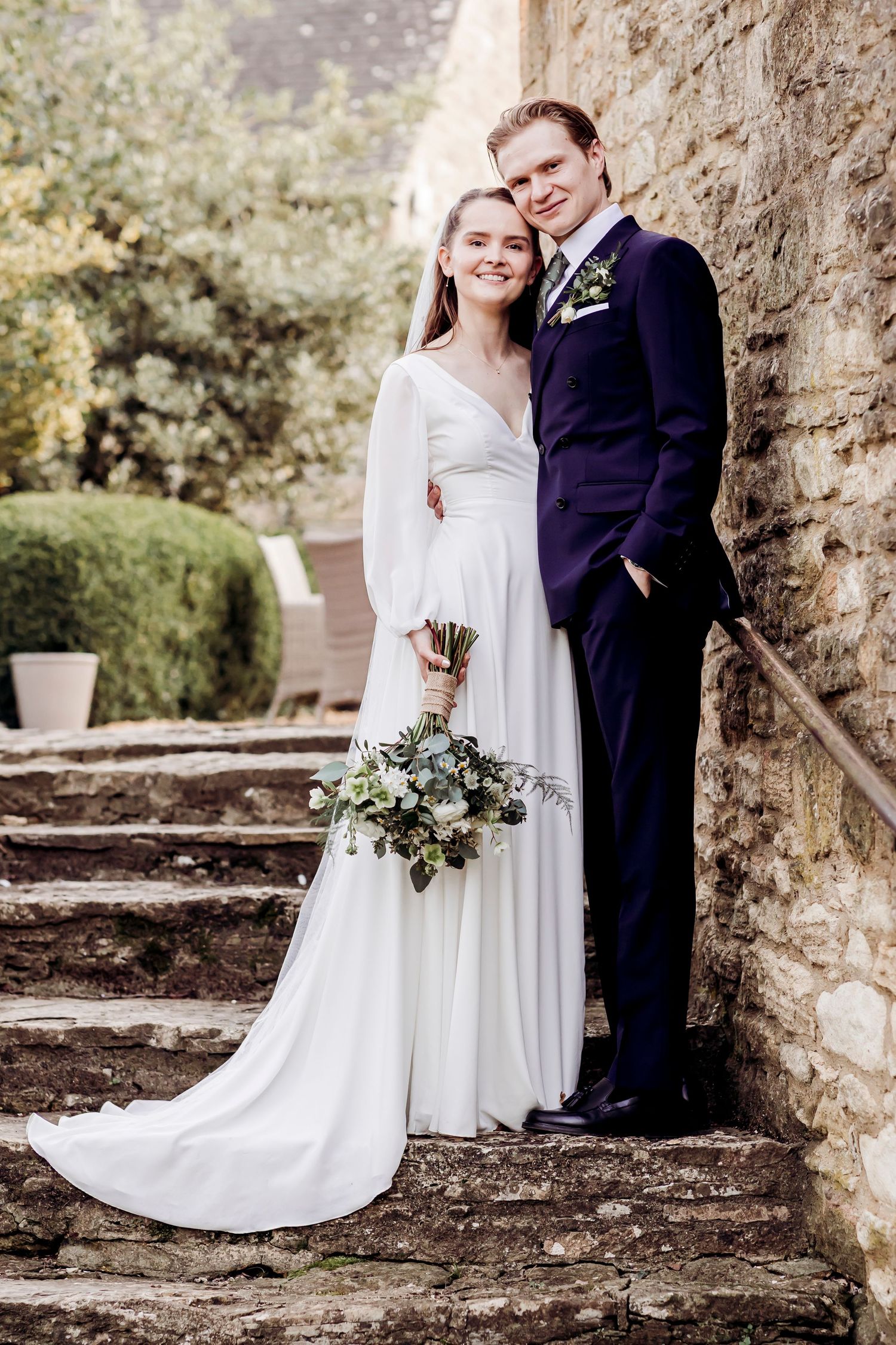 Bride in white gown and groom in navy suit posing on stone steps outdoors.