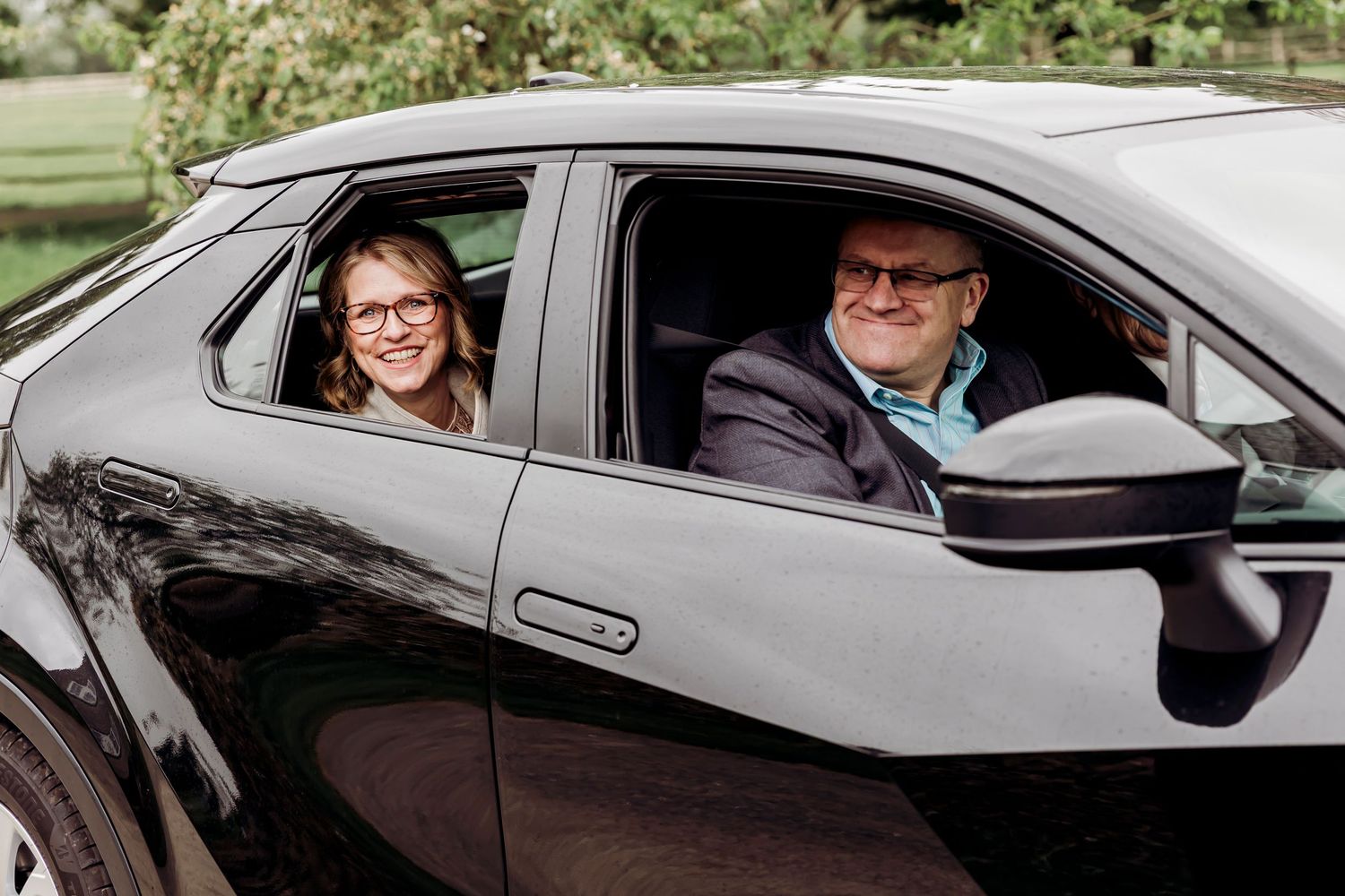 A smiling woman and older man pose together inside a sleek black car on a sunny day.