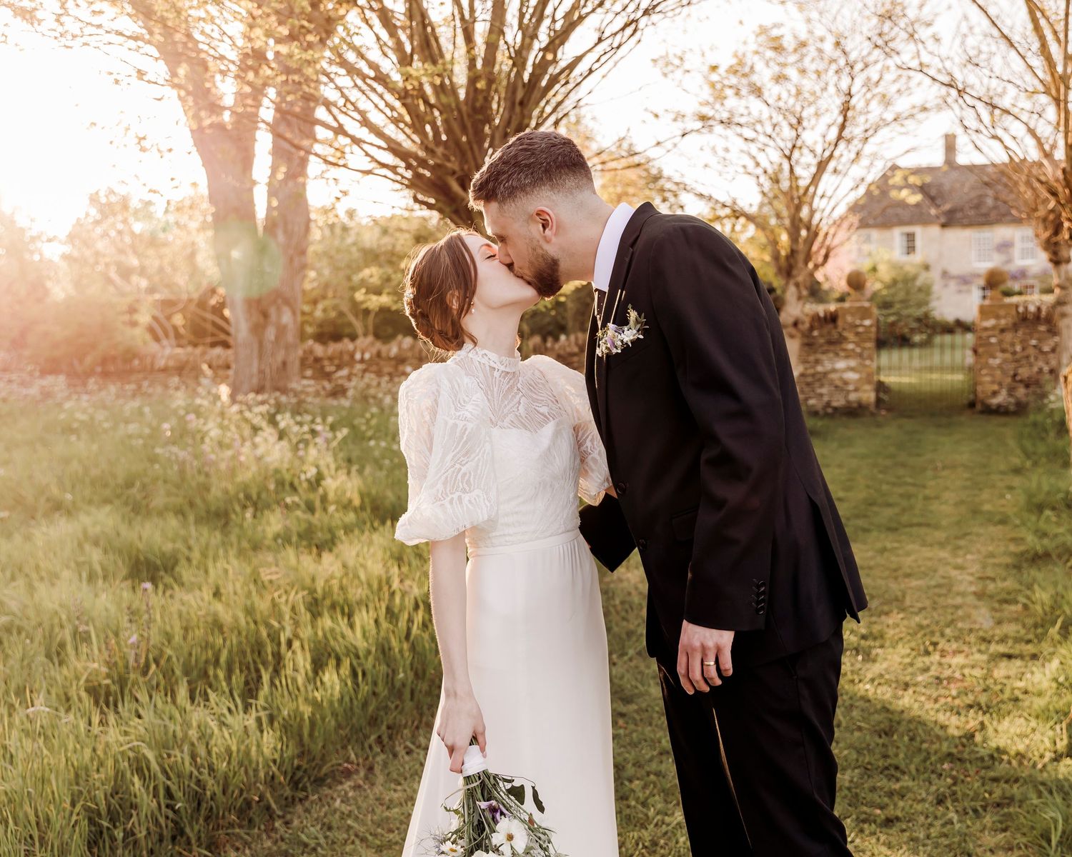 Bride and groom share a romantic kiss in a golden sunset garden, bride holding a white bouquet.