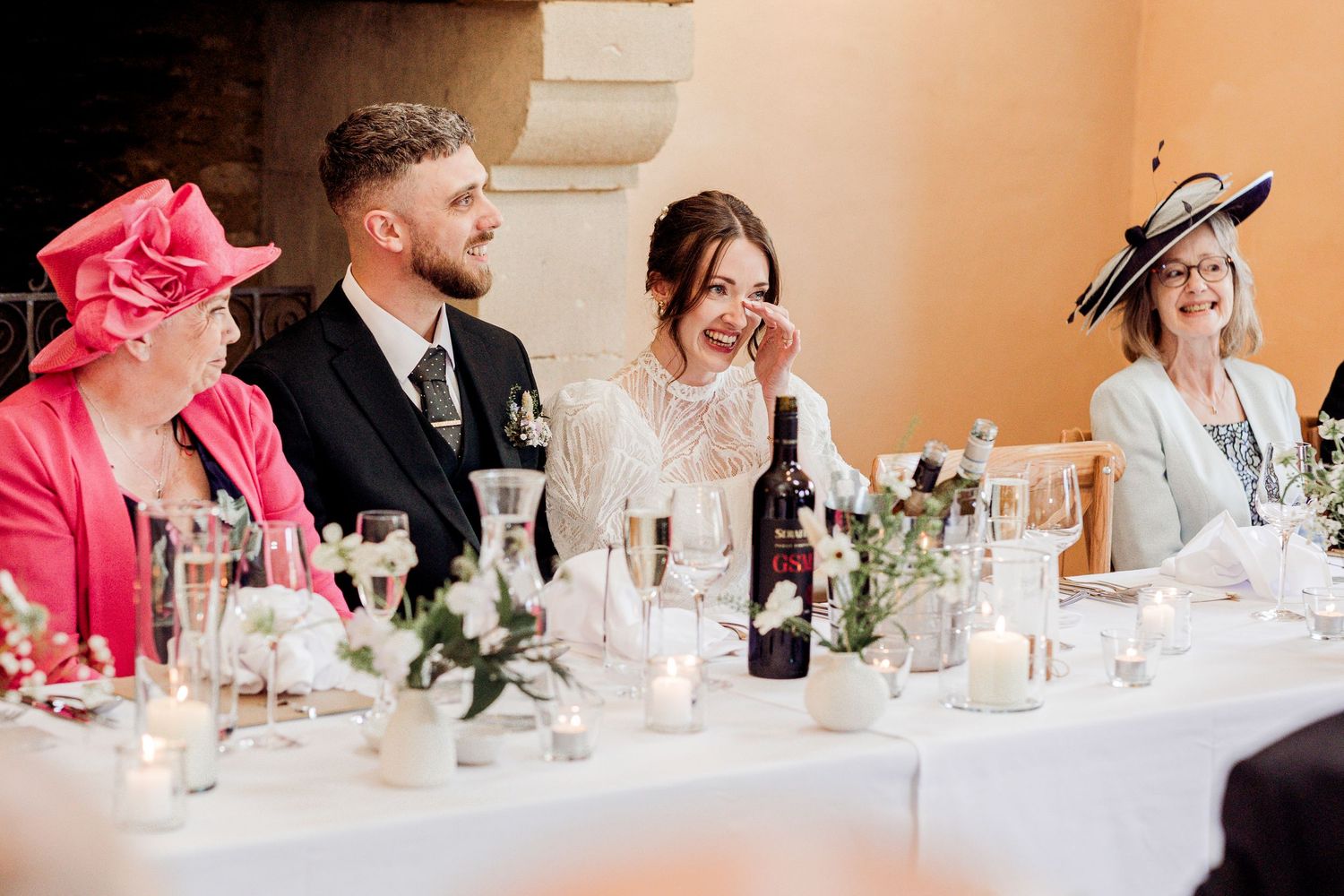 Bride and groom laugh at wedding reception head table surrounded by candles, flowers, and elegantly dressed guests.
