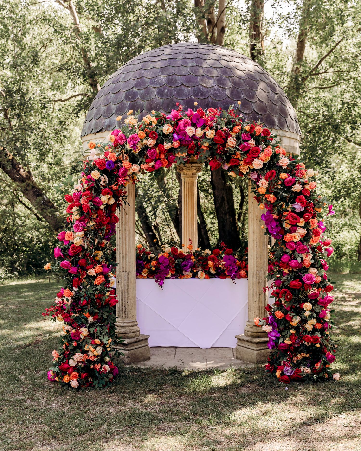 A stunning floral arch with vibrant pink, orange, and red roses framing a garden gazebo altar.