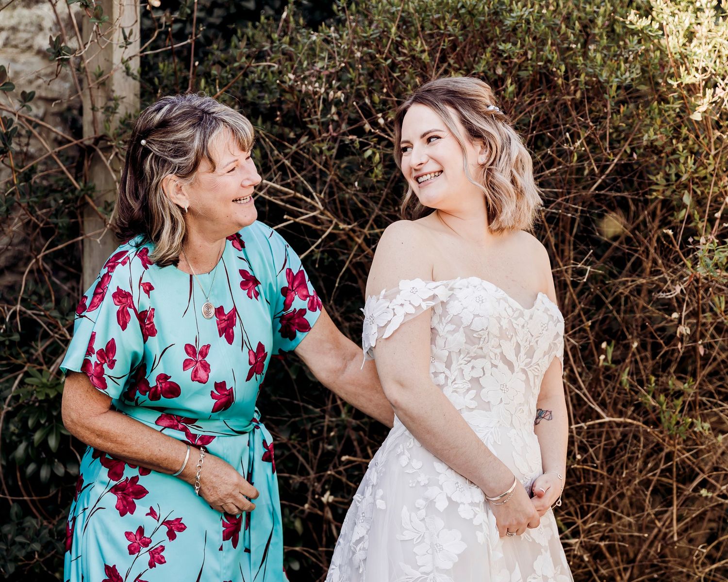Bride in floral lace wedding dress sharing a joyful moment with her mother outdoors.