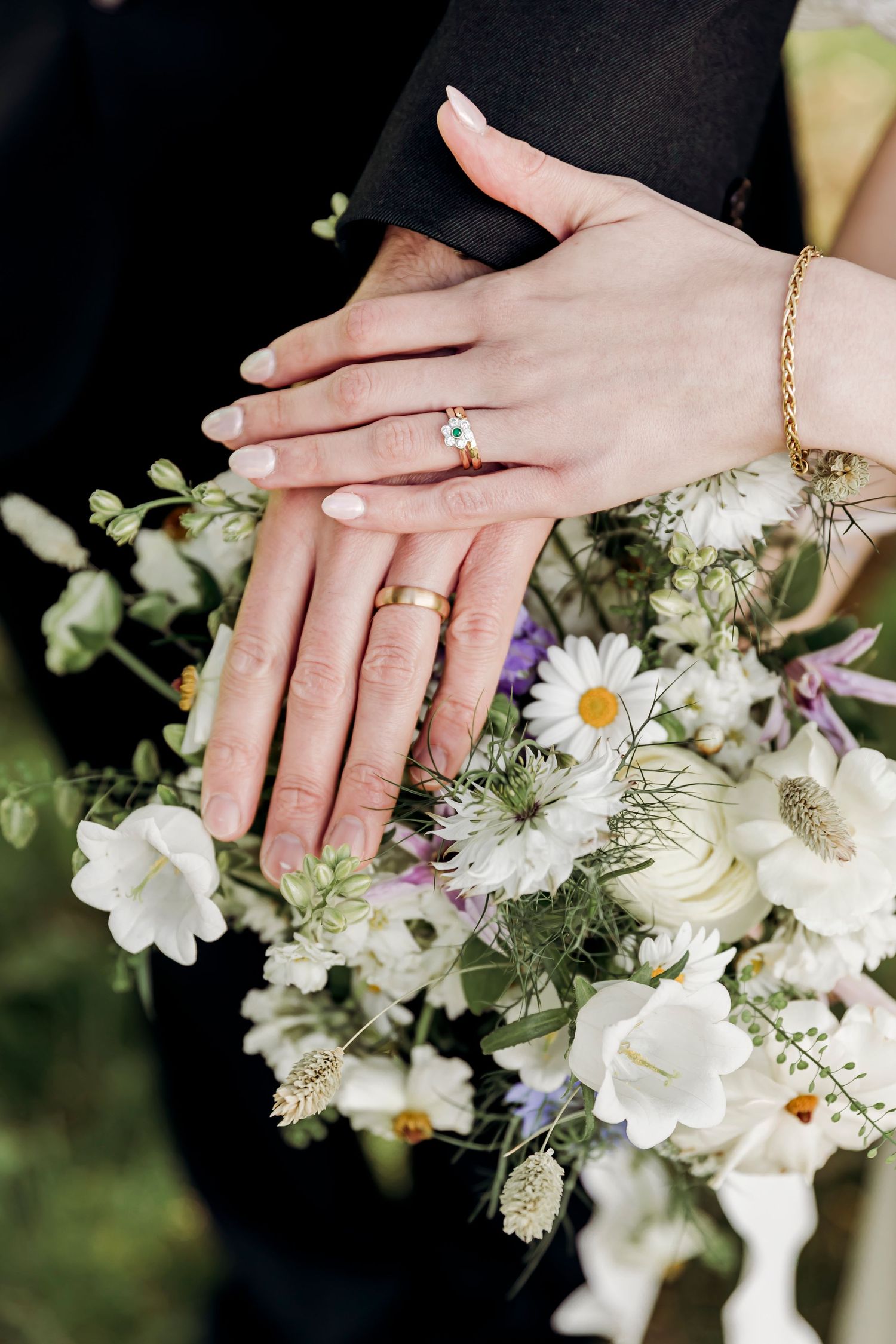 Newlyweds display gold wedding rings and engagement ring on hands resting over a white wildflower bridal bouquet.