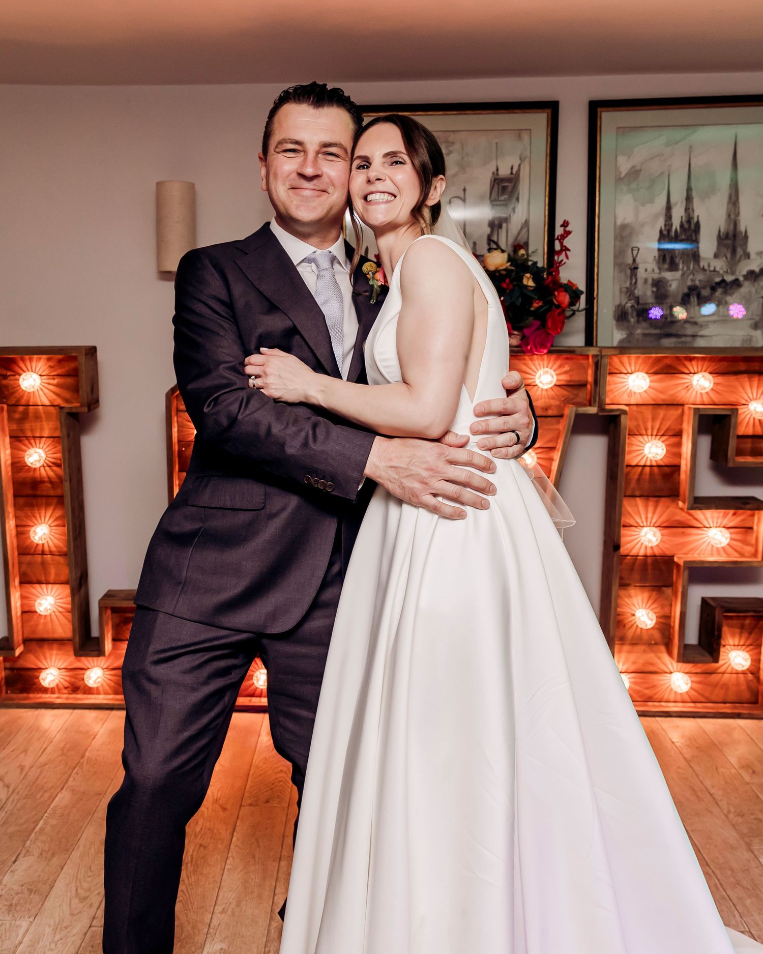 Bride and groom embracing in front of illuminated marquee letters at their wedding reception.