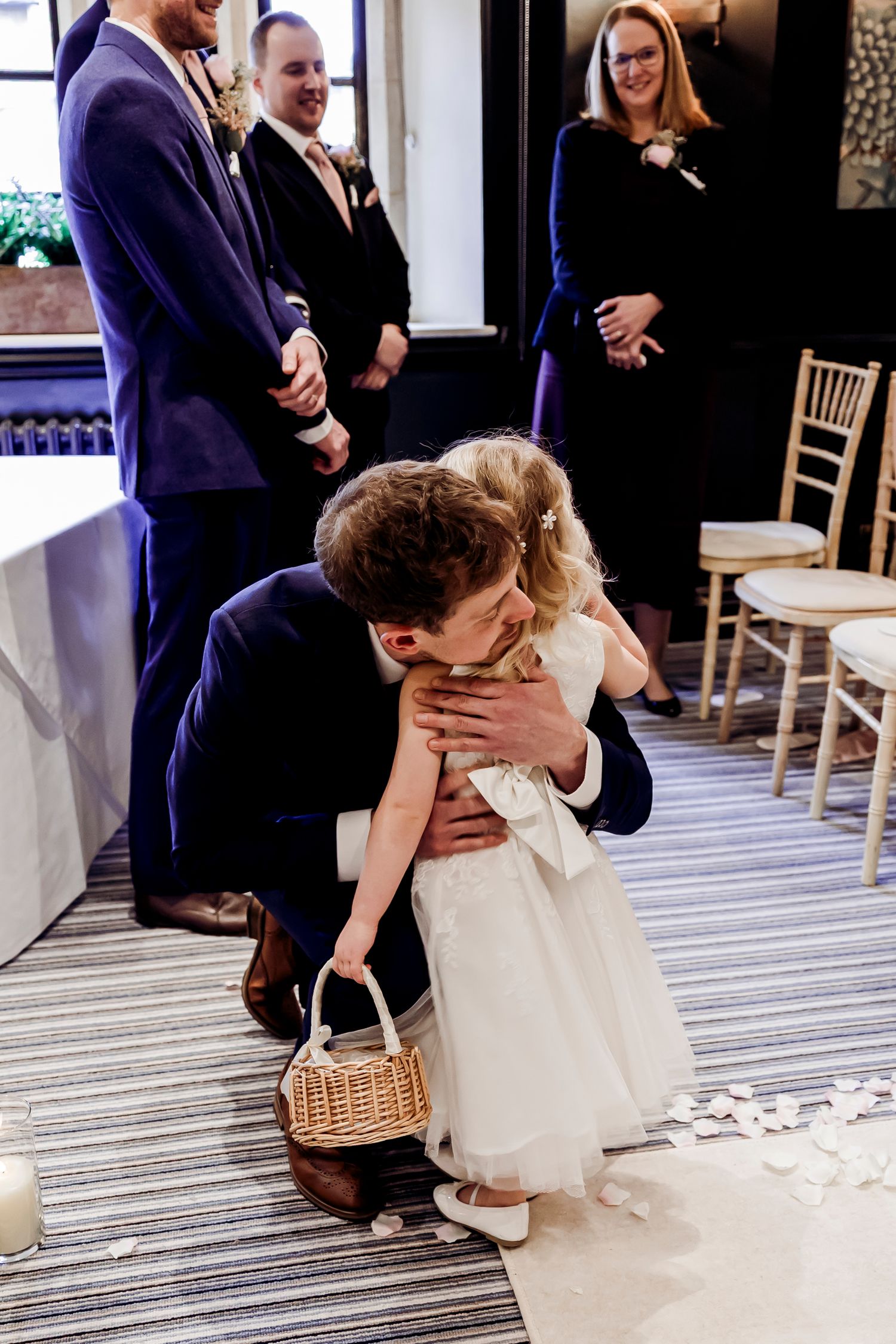 Groom kneeling to embrace flower girl in white dress during indoor wedding ceremony.