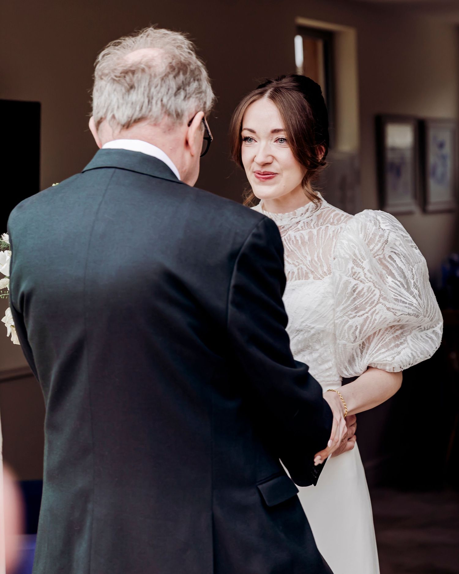 Bride in white lace gown holding hands with guest during wedding ceremony.