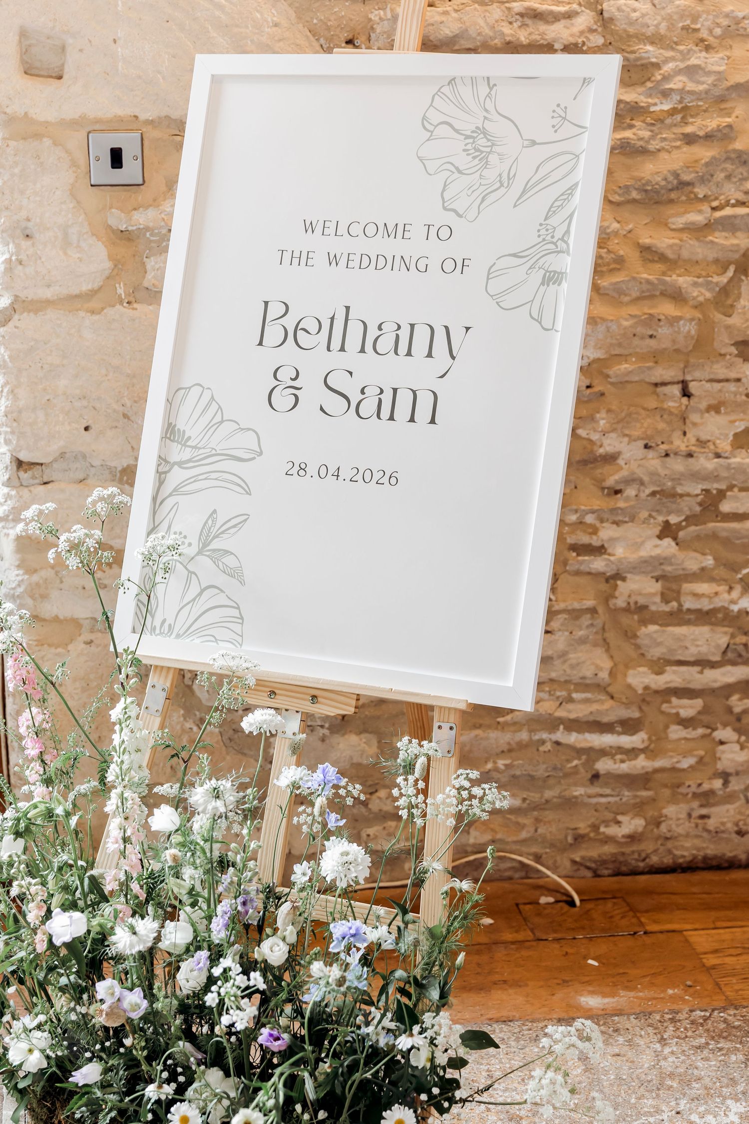 Elegant wedding welcome sign for Bethany and Sam on a wooden easel surrounded by wildflowers against a stone wall.