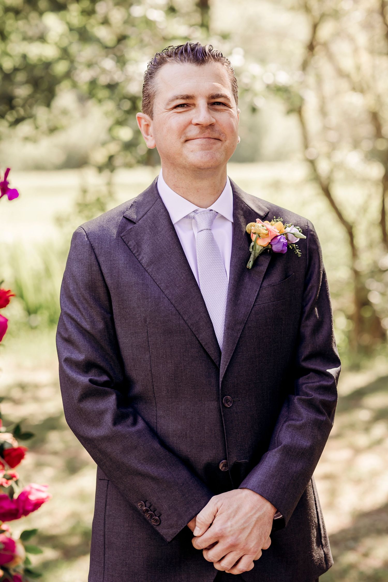 A smiling groom in a dark suit with a colorful boutonniere standing outdoors at a wedding.