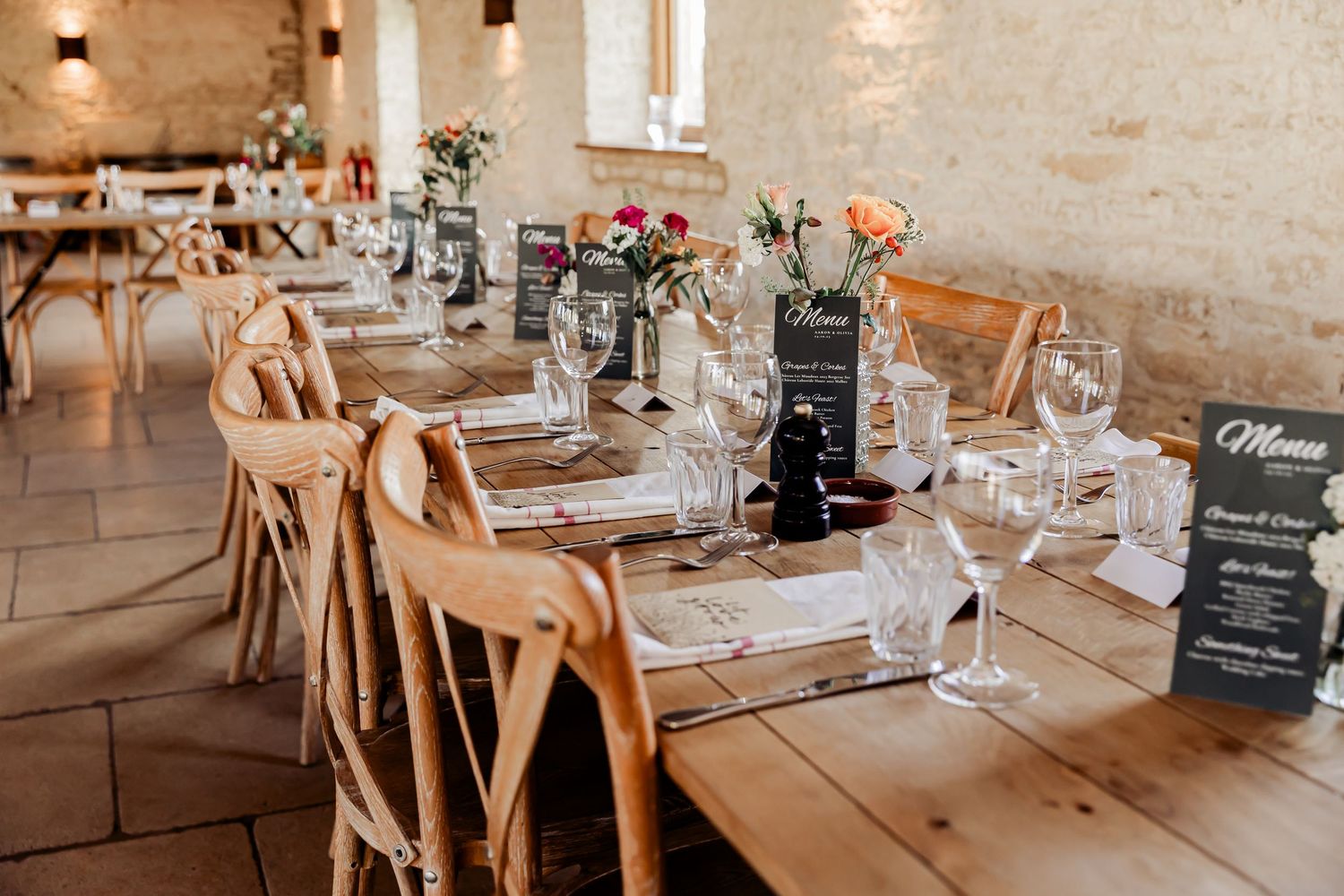 Elegant rustic wedding reception with long wooden tables, cross-back chairs, floral centerpieces, and menu cards.