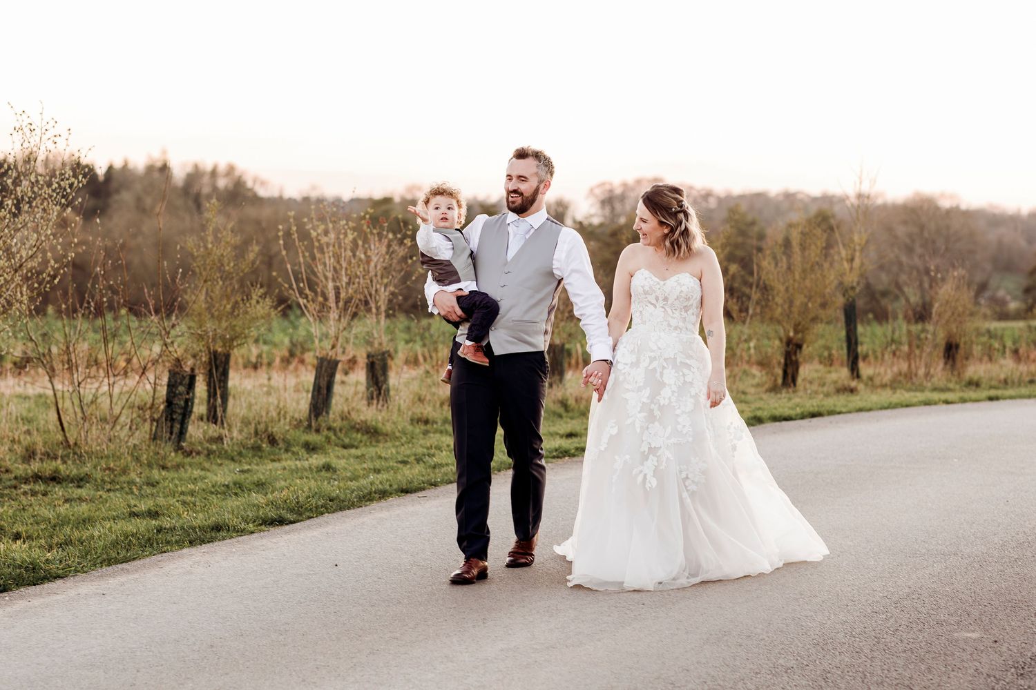 Bride and groom walking with young child on a scenic road at golden hour.
