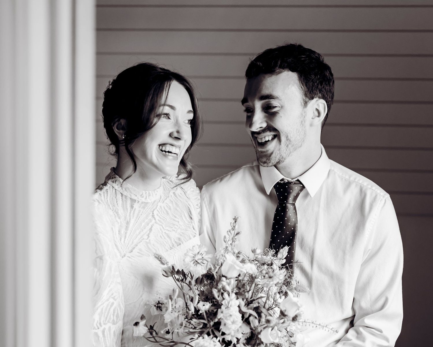 Black and white photo of a smiling bride and groom sharing a joyful moment, bride holding a floral bouquet.