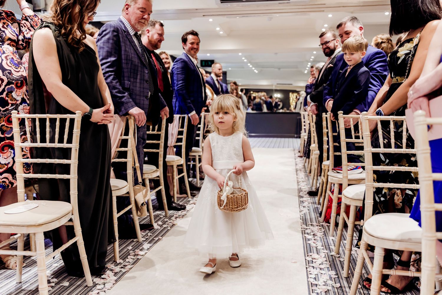 Adorable flower girl in white dress walking down wedding aisle carrying wicker basket.