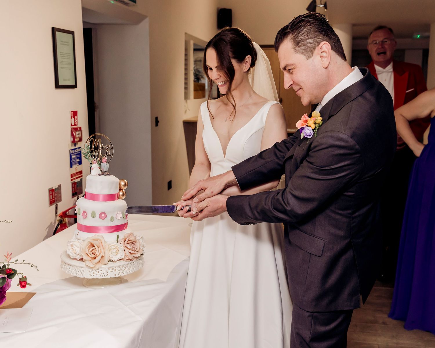 Bride and groom in elegant attire cut tiered white wedding cake decorated with pink ribbon and flowers.
