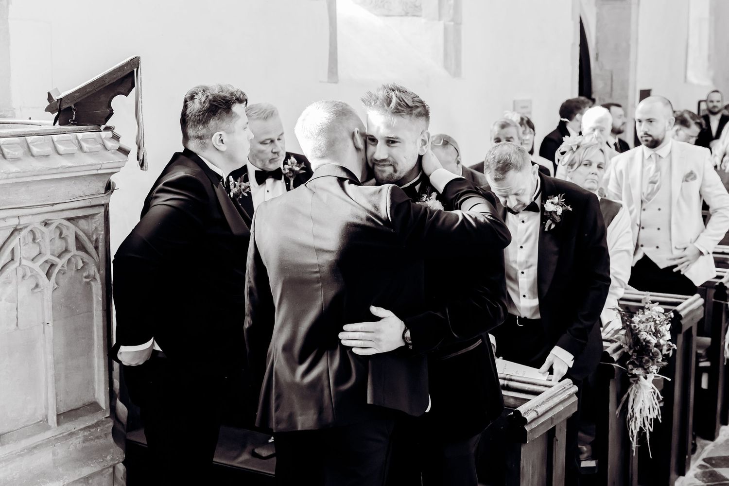 Black and white photo of groom sharing an emotional hug with guest inside a church ceremony.