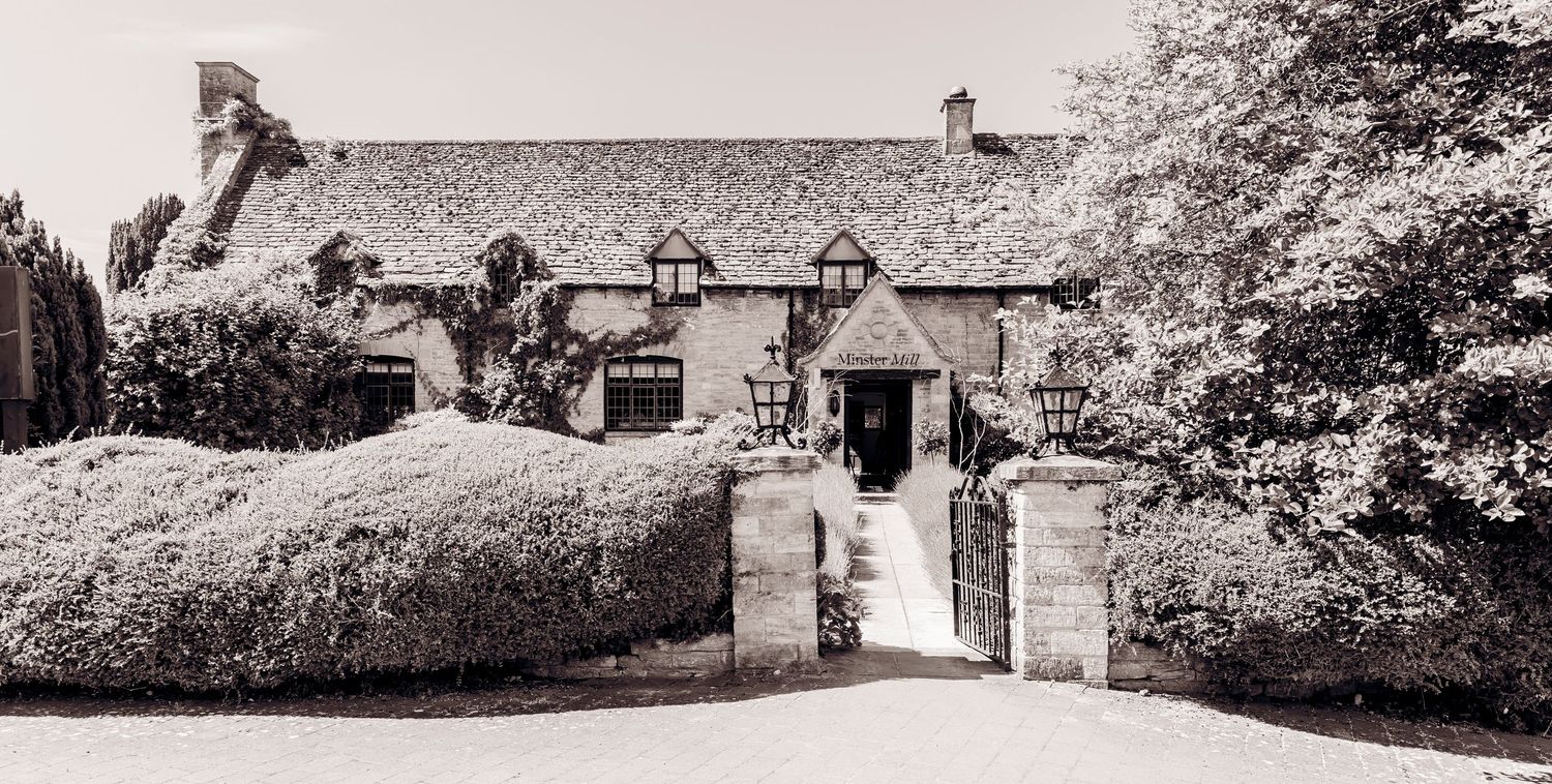 Black and white photo of a charming stone country house with manicured hedges and gated entrance.