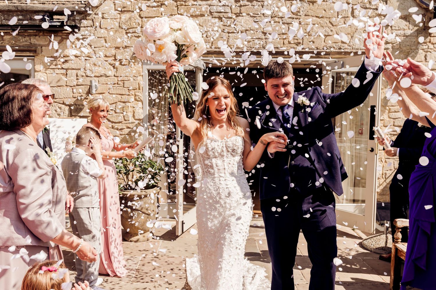 Bride and groom celebrate with confetti shower outside stone venue, guests cheering around them on their wedding day.
