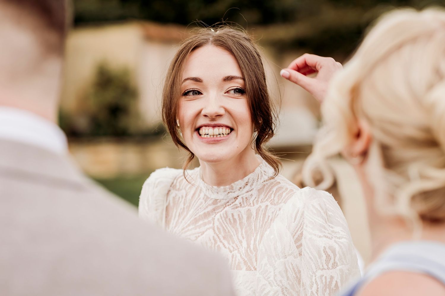 Smiling bride in white lace high-neck gown laughing with guests at outdoor wedding celebration.