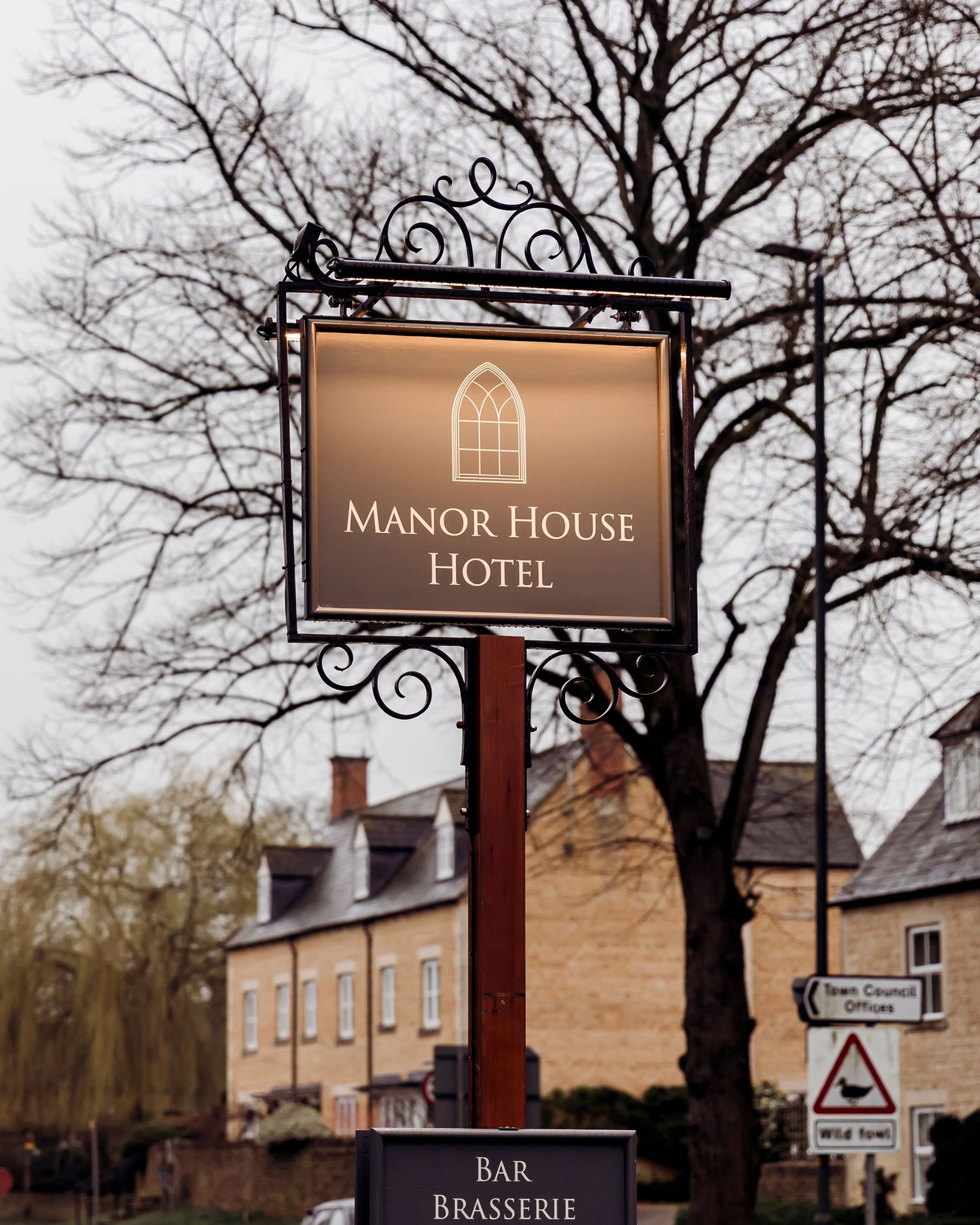 Manor House Hotel sign on an ornate iron post surrounded by bare winter trees.