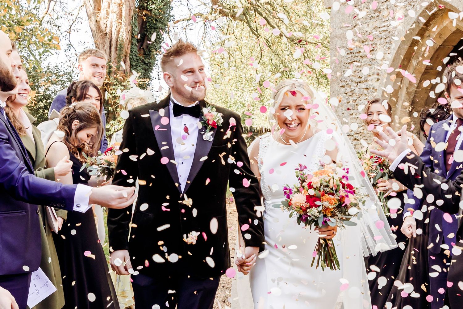 Bride and groom walk through a shower of pink and white confetti thrown by cheering wedding guests.