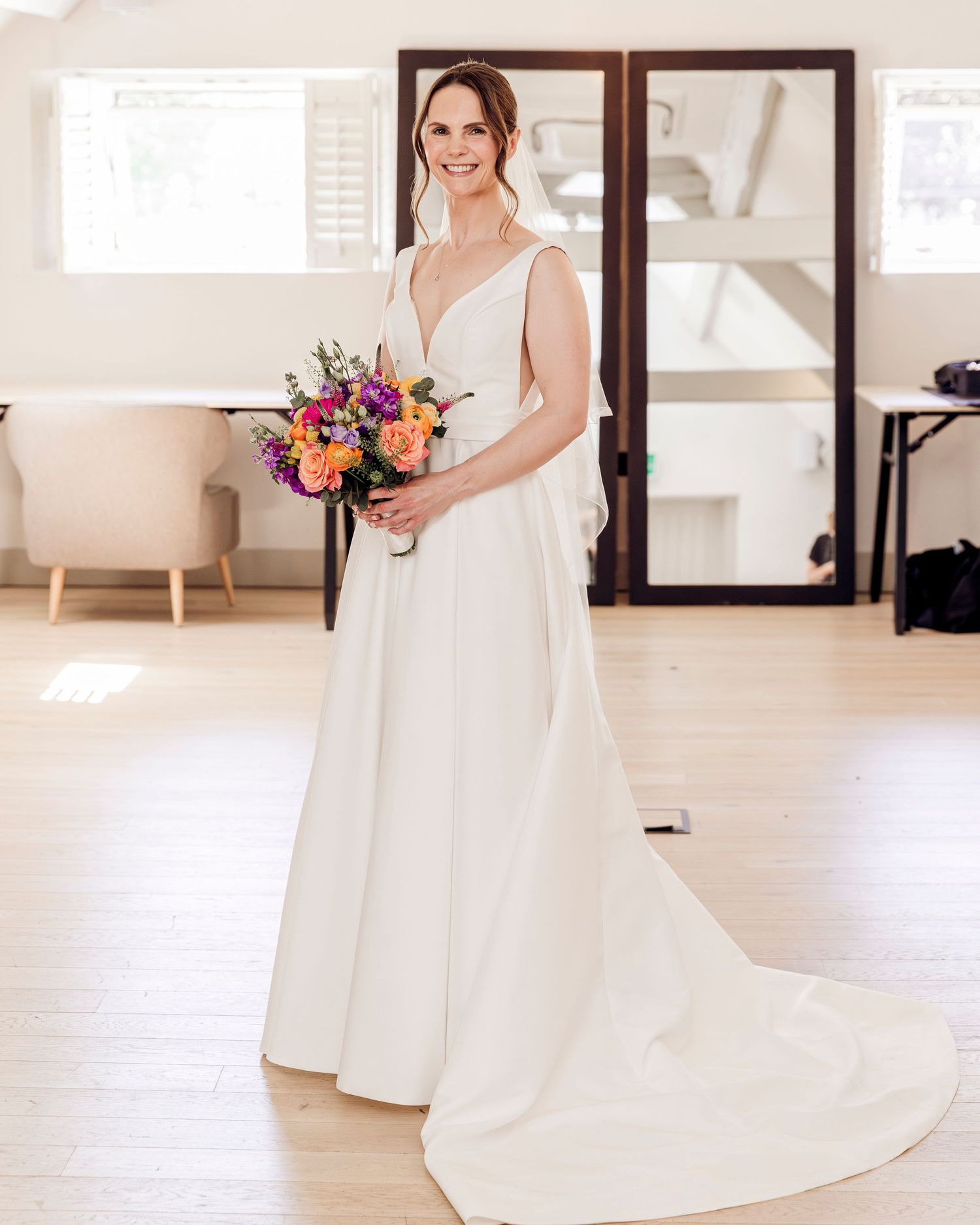 Bride in elegant white V-neck wedding gown holding colorful bouquet of purple and orange flowers in bright room.