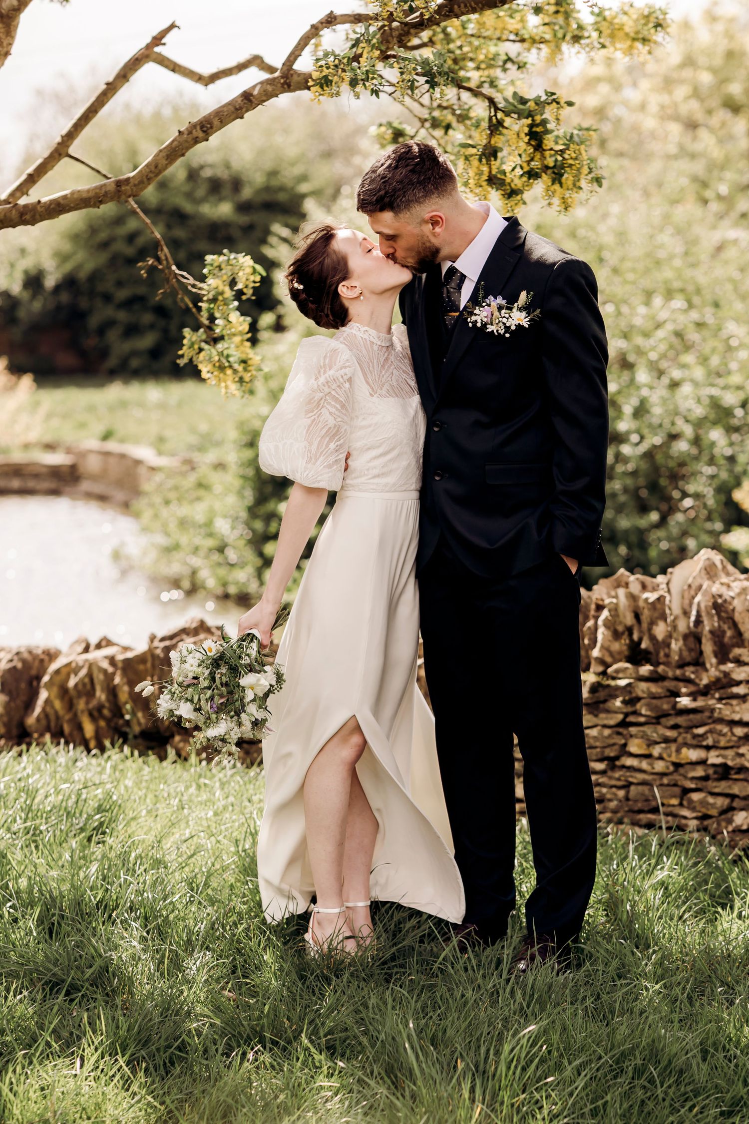 Bride in white lace dress and groom in dark suit share a kiss in a lush green garden setting.