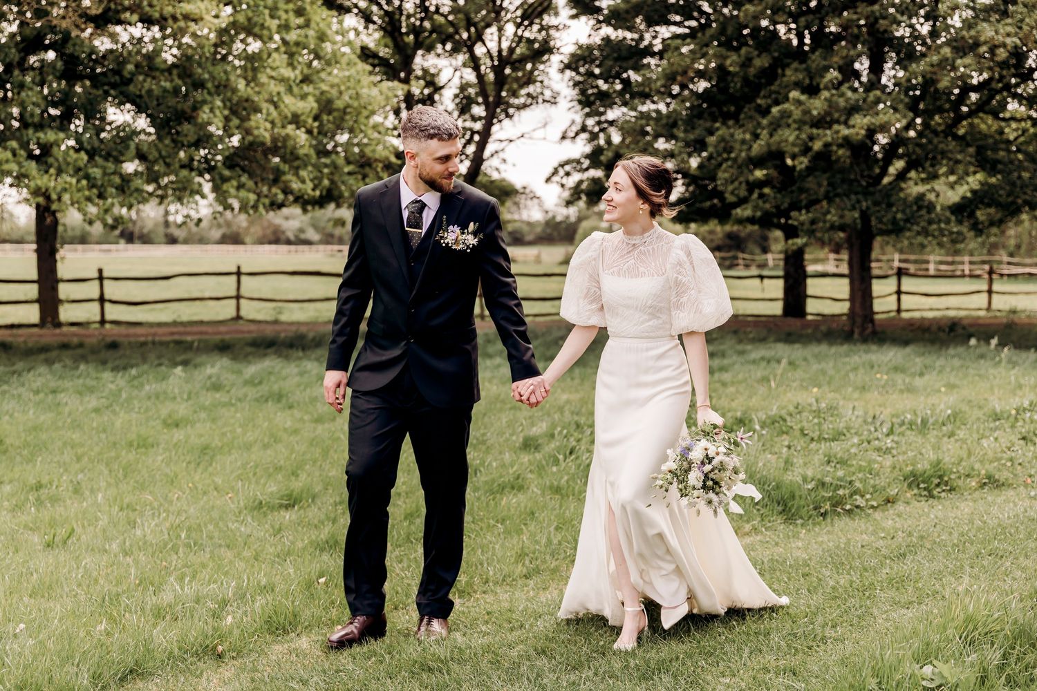 Bride and groom walk hand-in-hand through a green meadow, bride holding a white bouquet.
