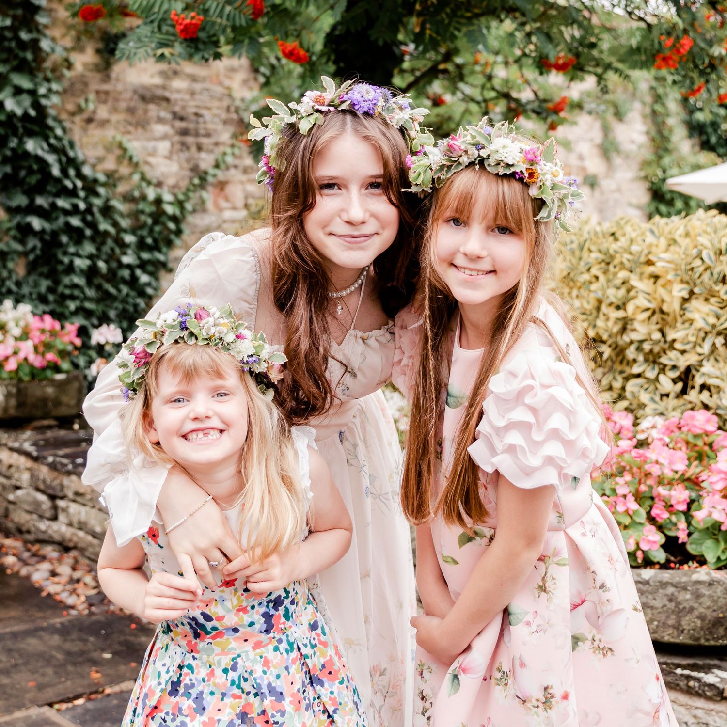 Three girls in floral crowns and dresses pose together in a garden with colorful blooms.