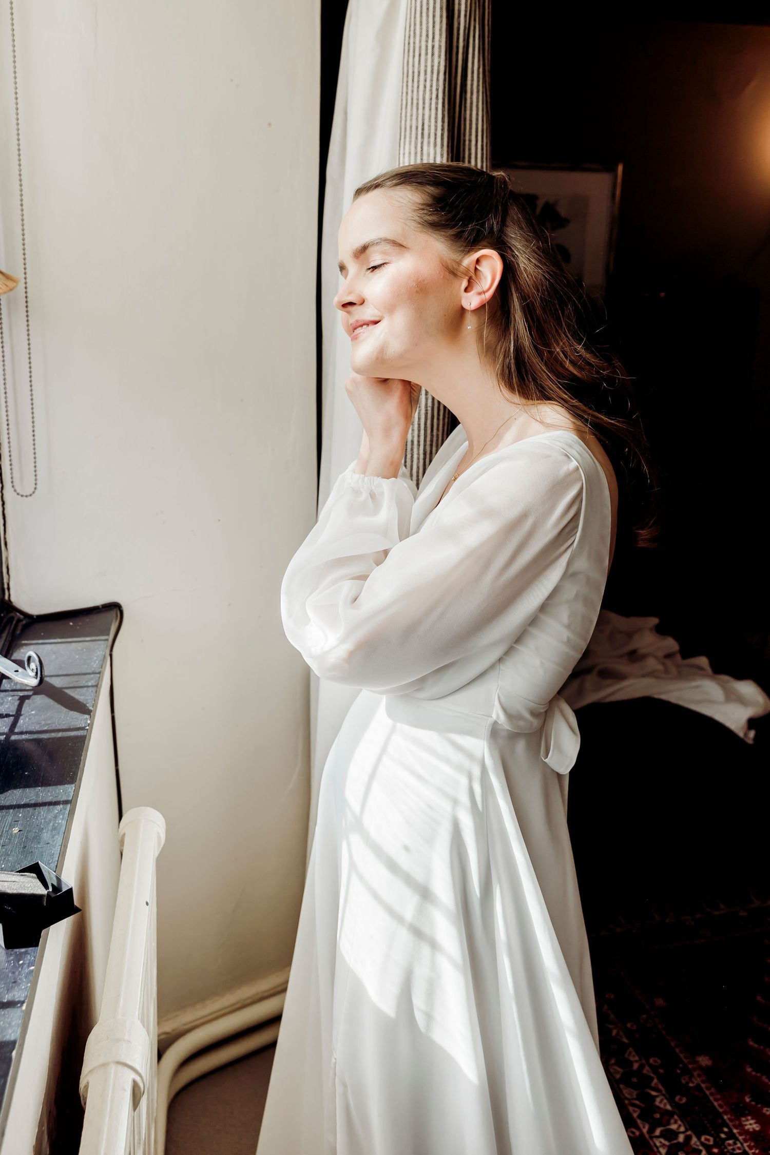 Beautiful bride in a white long-sleeve wedding dress smiling with eyes closed by a window.