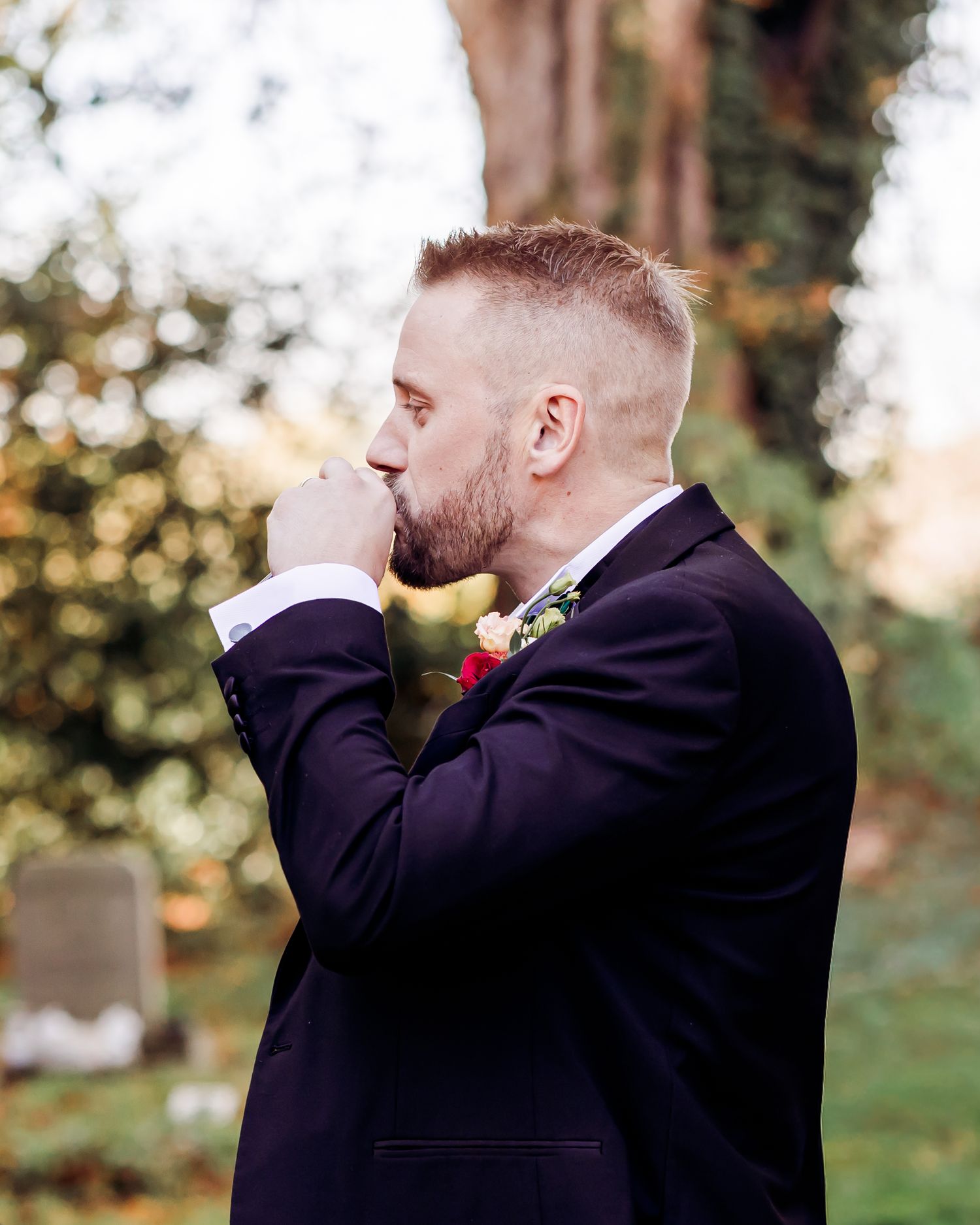 Groom in dark suit with floral boutonniere standing thoughtfully outdoors in black and white photo.