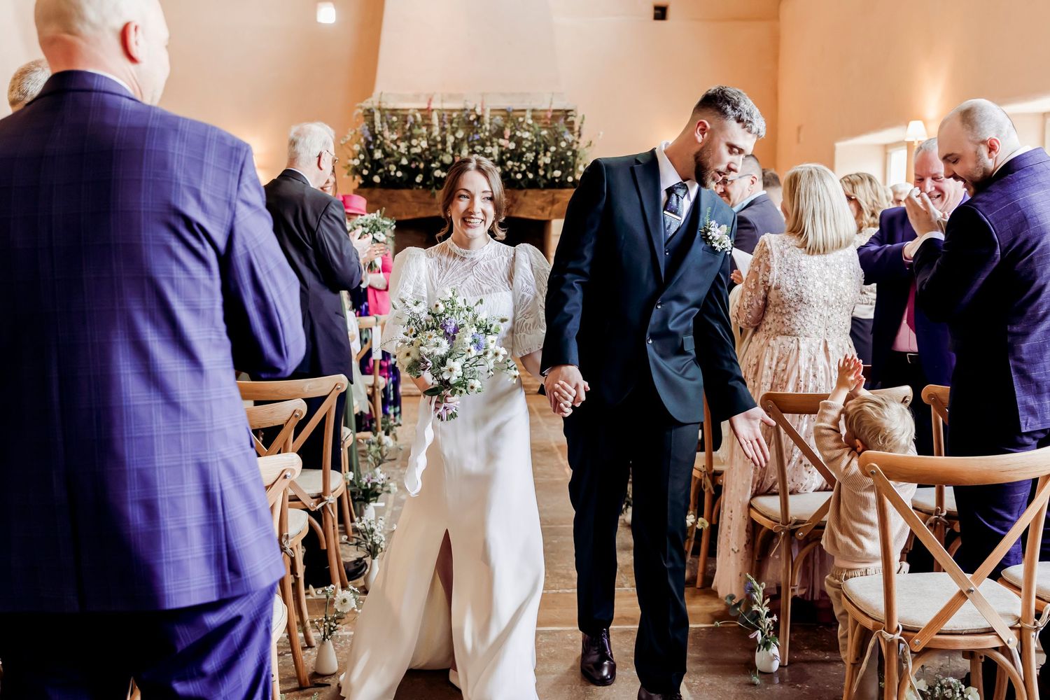 Newlywed couple walks hand-in-hand down the aisle after wedding ceremony, bride in white jumpsuit holding bouquet.