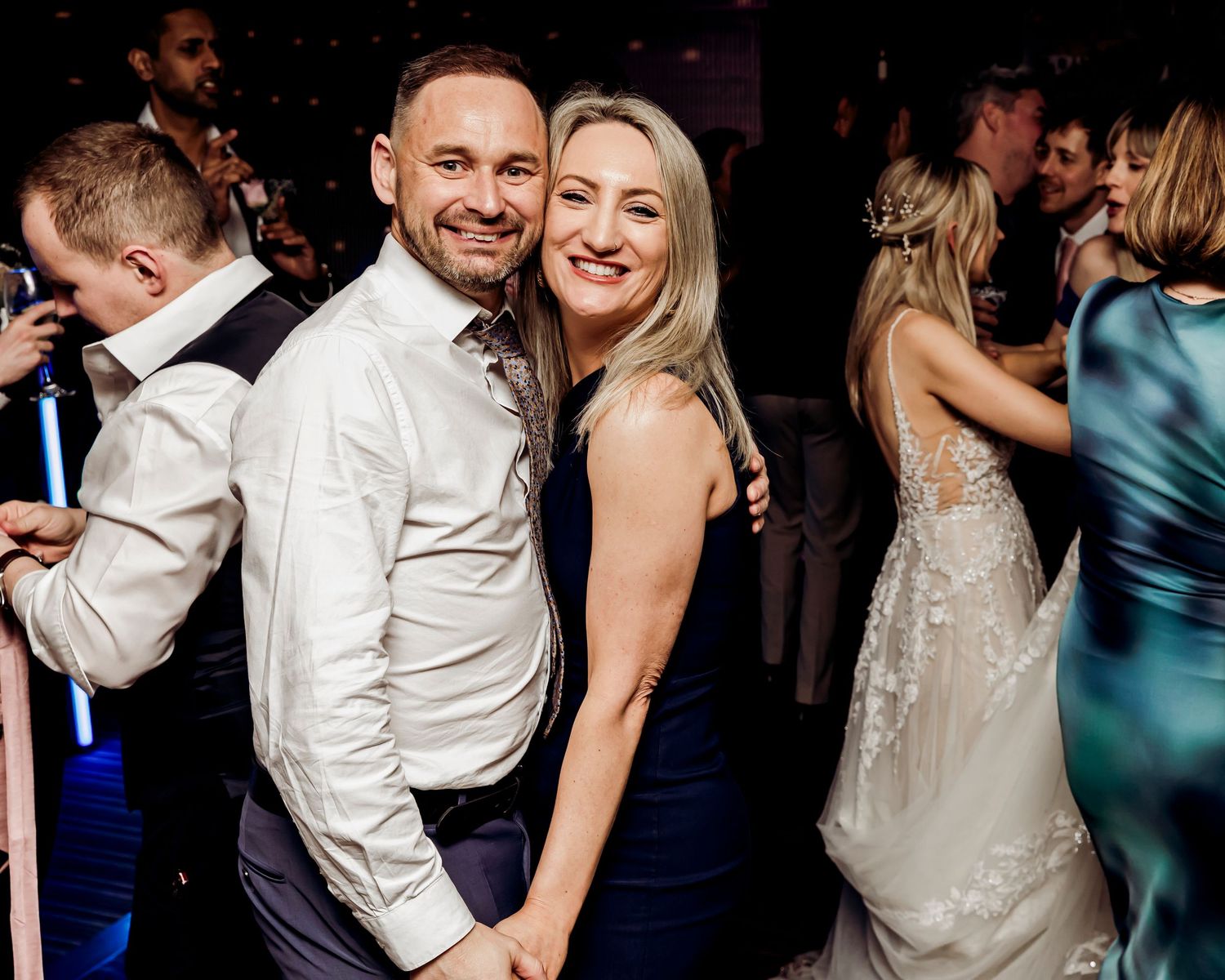 Happy couple poses together on the wedding dance floor, bride in a lace gown and groom in a white shirt.