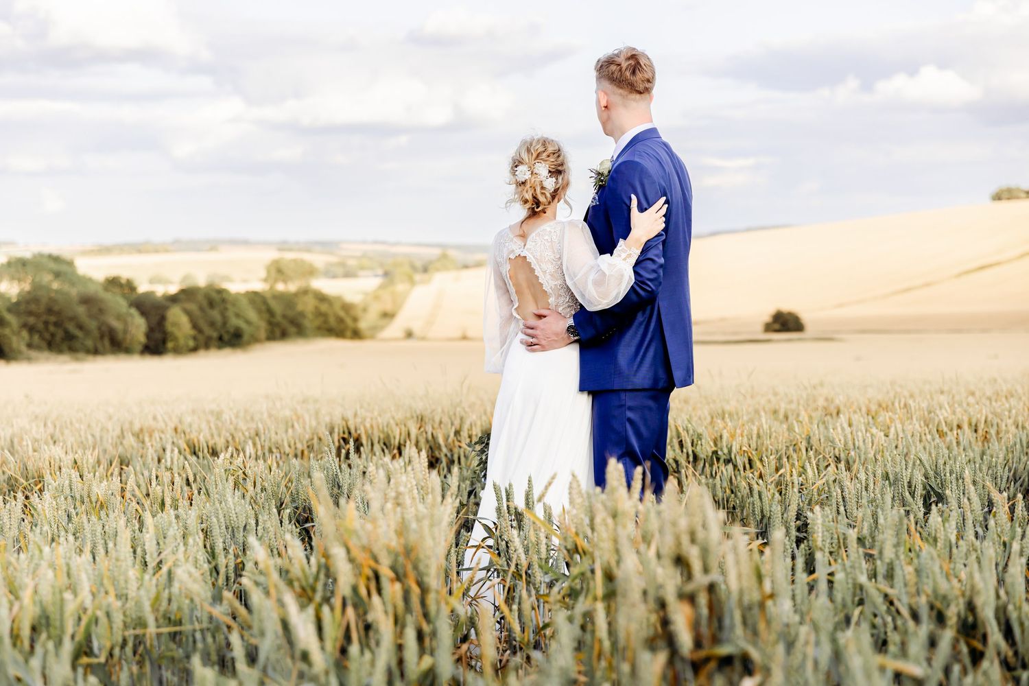 Bride and groom embrace romantically in a golden wheat field under a cloudy sky on their wedding day.