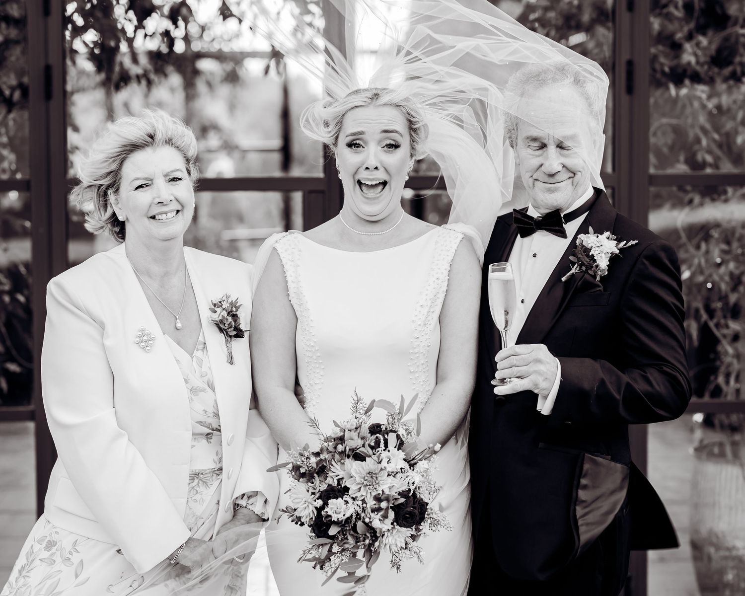 Black and white photo of laughing bride with bouquet flanked by proud parents at wedding celebration.