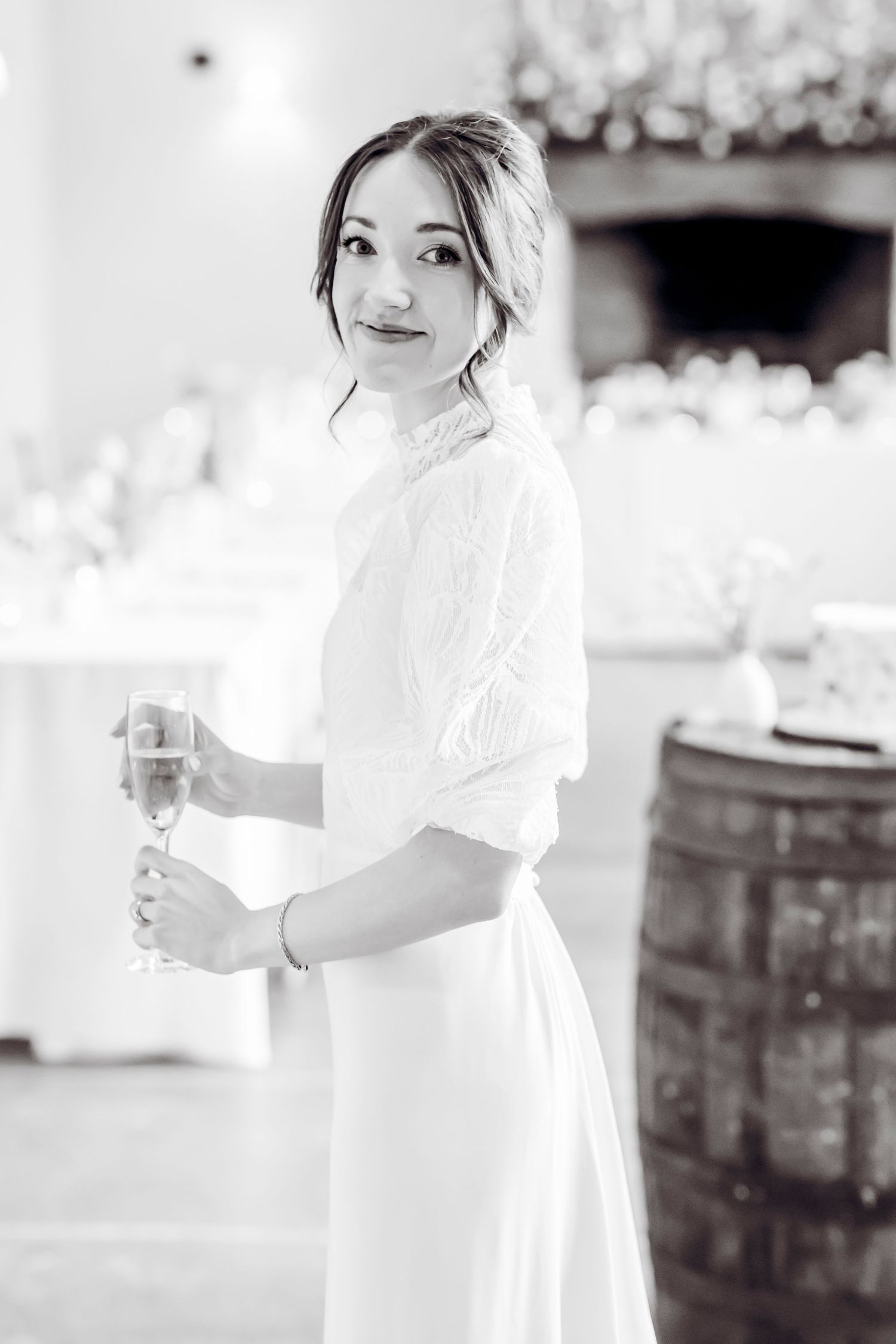 Black and white photo of bride in white lace dress holding champagne glass in decorated wedding venue.