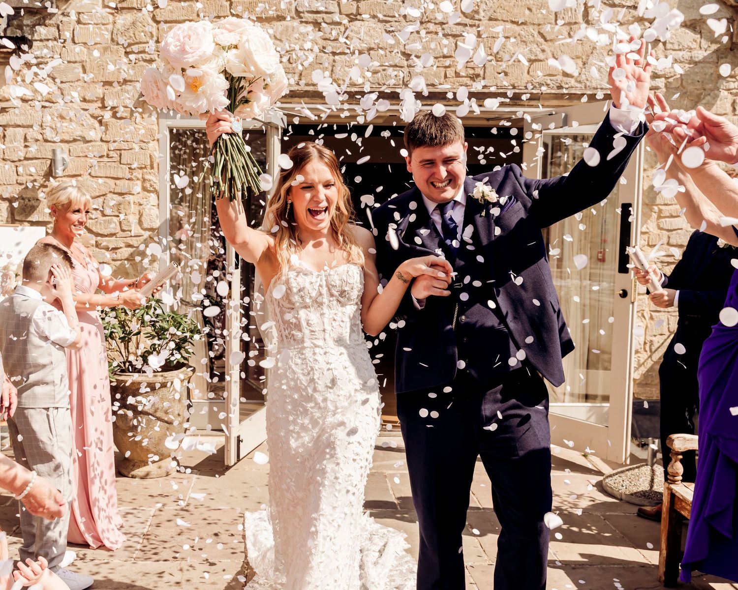 Happy newlyweds walk through confetti celebration outside rustic stone venue on sunny wedding day.