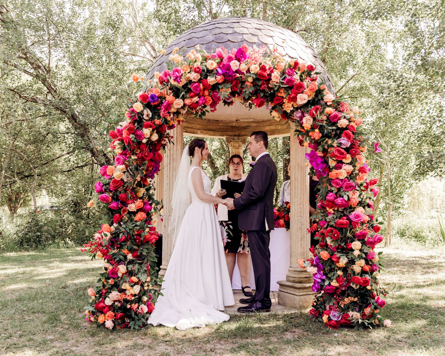 Bride and groom exchange vows under stunning colorful floral arch with pink and orange roses outdoors.
