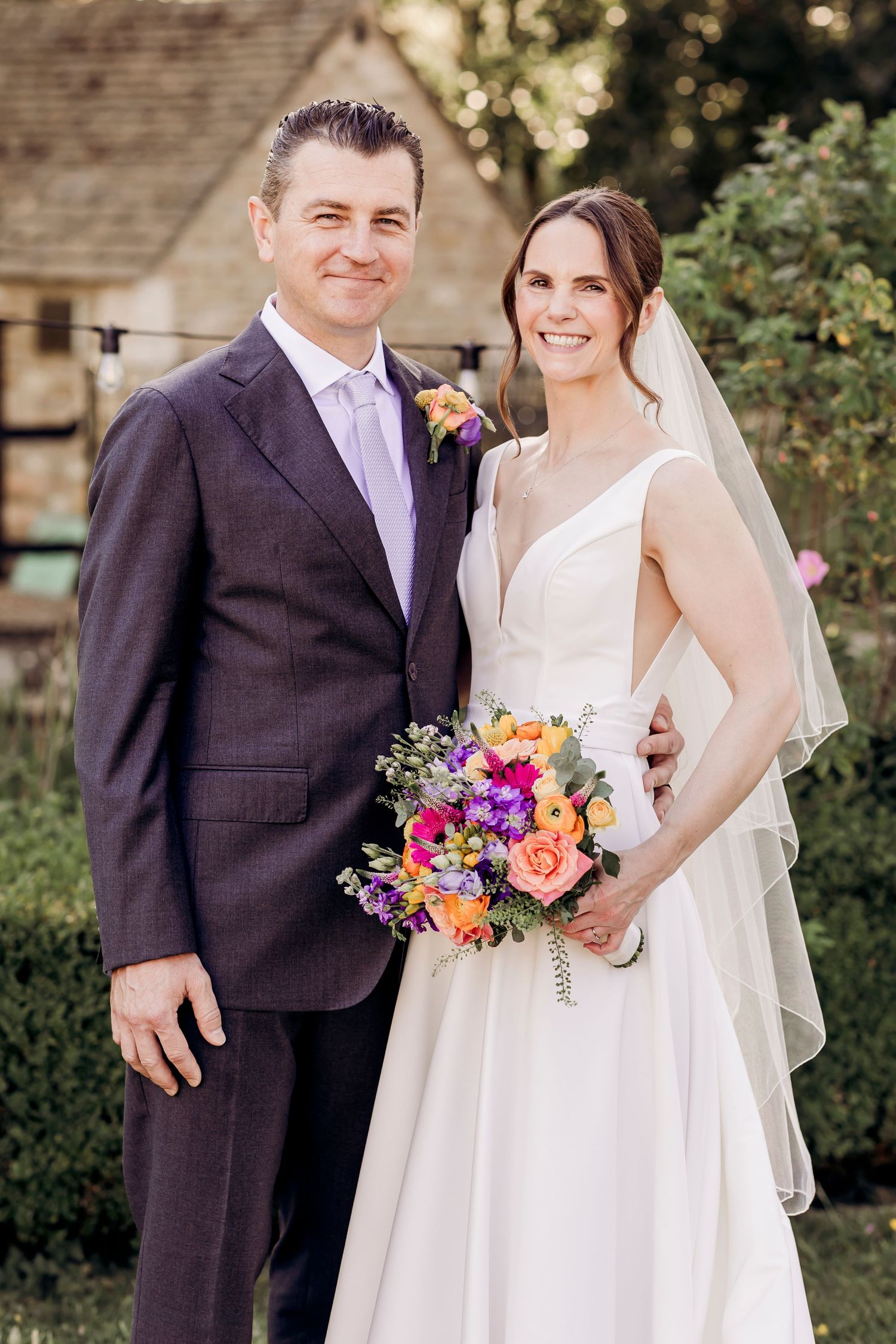 Bride and groom posing together outdoors, bride holding colorful bouquet in white gown with veil, groom in gray suit.