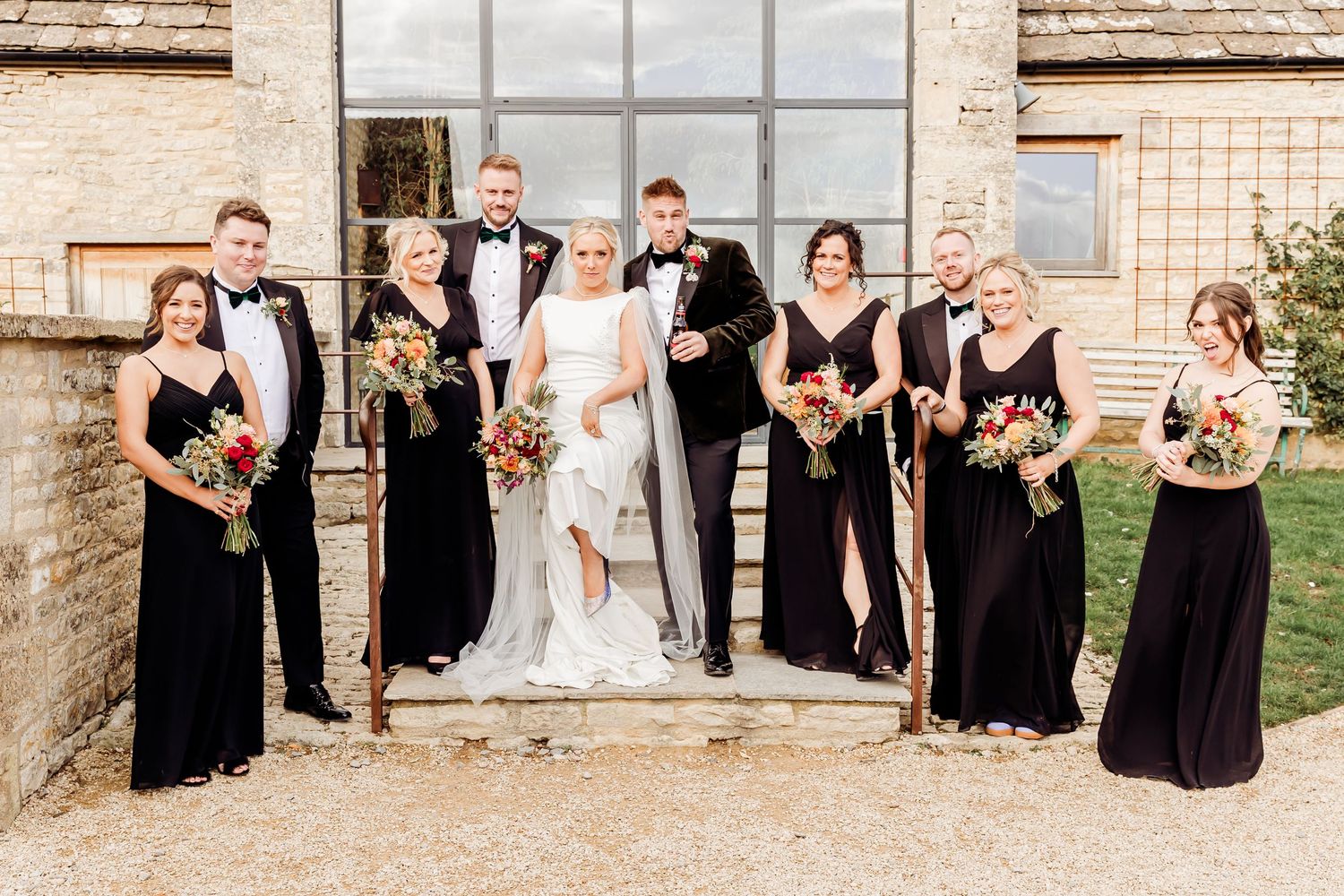 Wedding party of bride, groom, groomsmen, and bridesmaids in black attire posing outside stone venue.