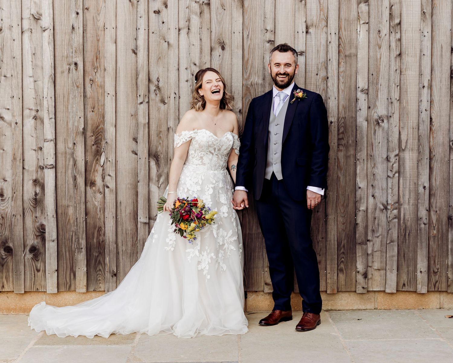 Happy bride and groom laugh together holding hands in front of rustic wooden barn wall, bride holding vibrant bouquet.